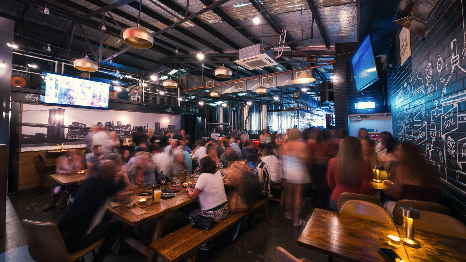 Crowded brewery beerhall with guests enjoying drinks in an industrial warehouse space.