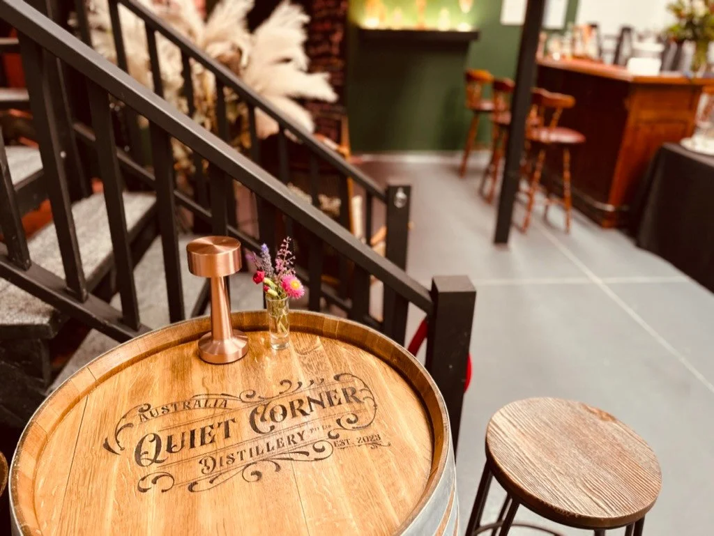 A wooden table with the words 'Quiet Corner' and 'Distillery' engraved on it, along with a small flower vase and a copper-colored lamp. A stool is next to the table, inside a cozy, rustic setting.
