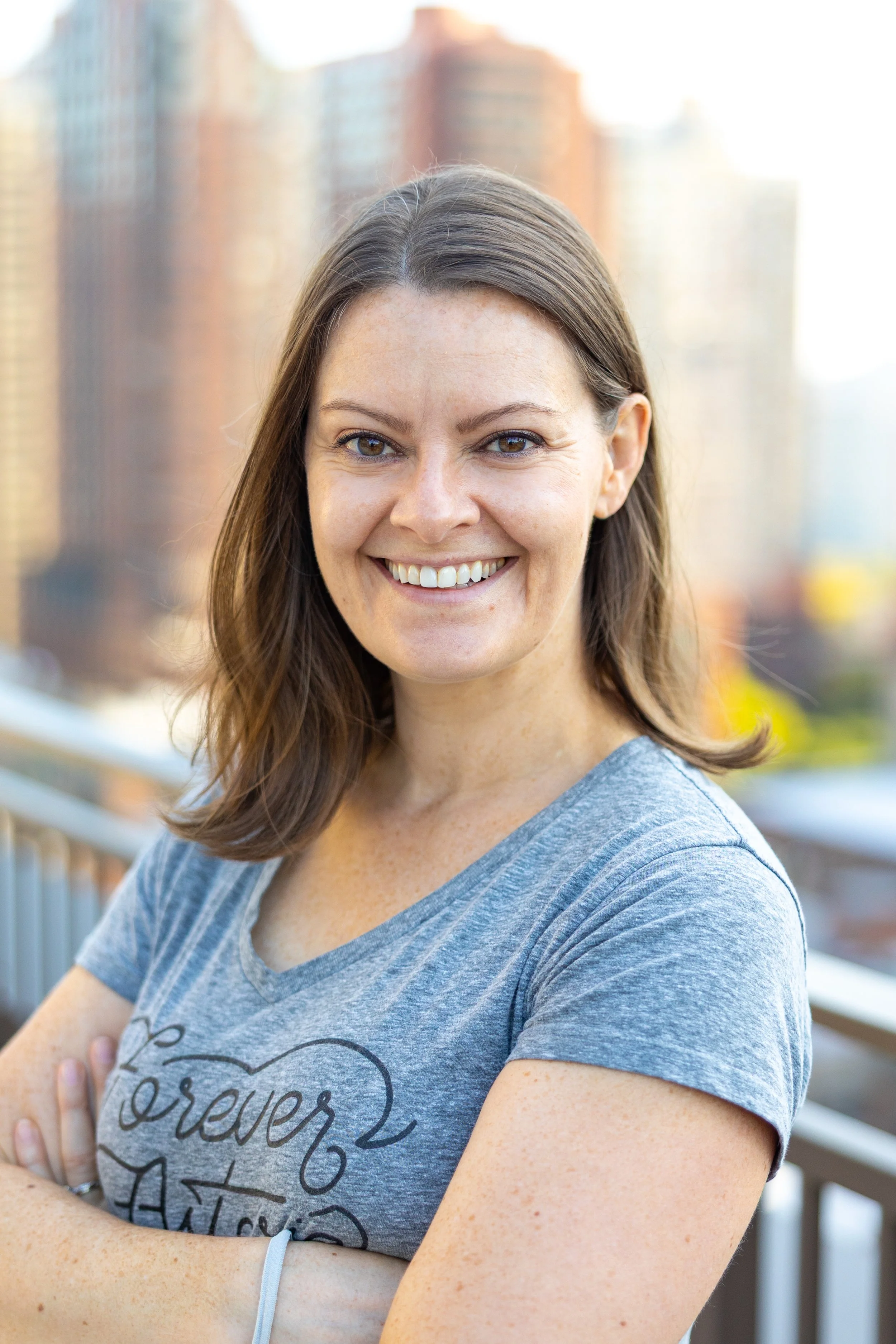 A woman with shoulder-length brown hair smiling, standing outdoors on a balcony with city buildings in the background, wearing a gray t-shirt.