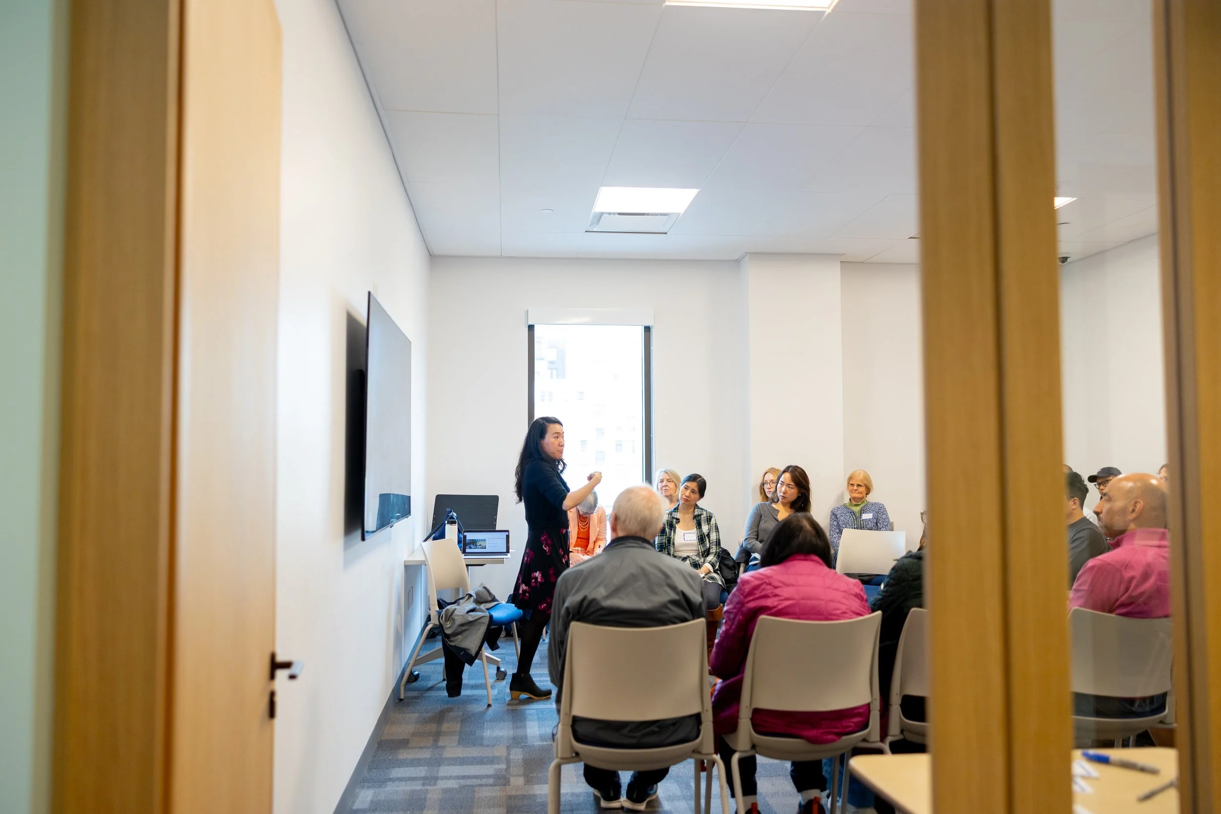 A woman standing and speaking to a group of seated people in a conference room, viewed through a doorway.