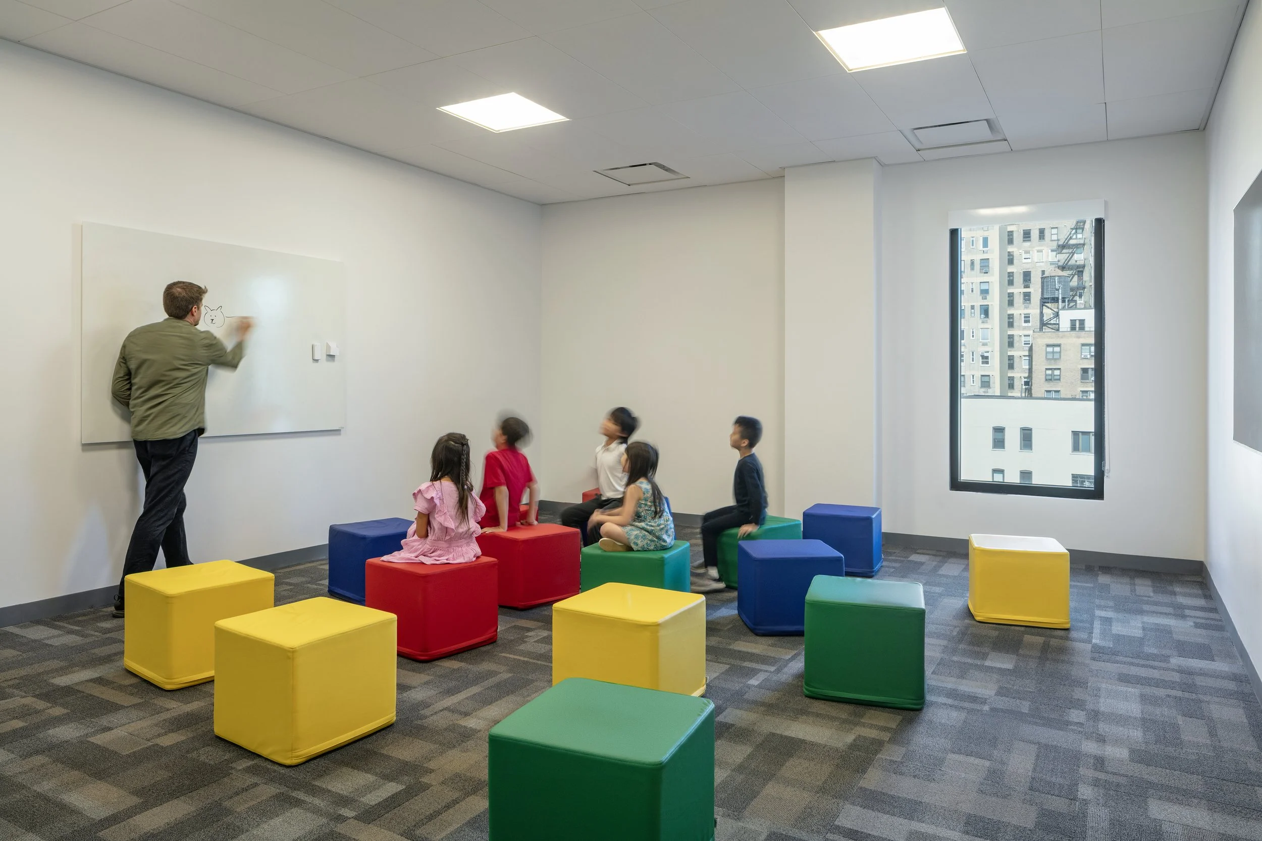 A teacher drawing on a whiteboard with a group of five children seated on colorful cushioned blocks in a classroom with a large window showing city buildings.