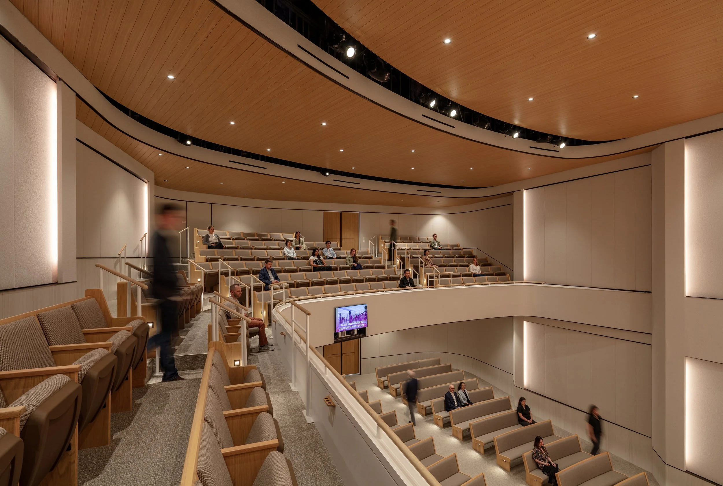 Interior of a modern theater or auditorium with multiple rows of beige seats, some occupied by people, and a wooden ceiling with recessed lighting.