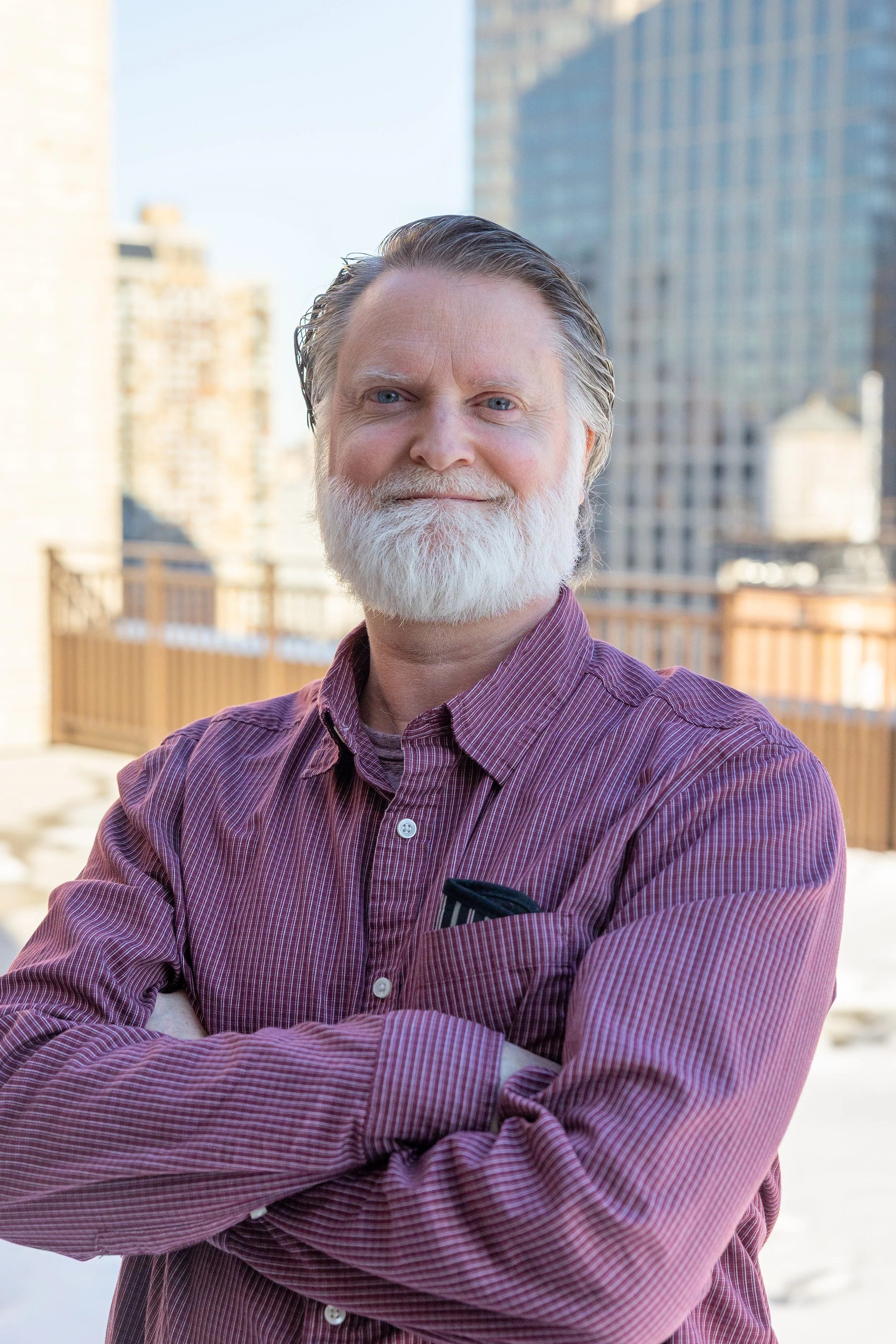 A senior man with a white beard and gray hair standing outdoors in an urban setting, wearing a purple checked shirt with arms crossed and a city skyline in the background.