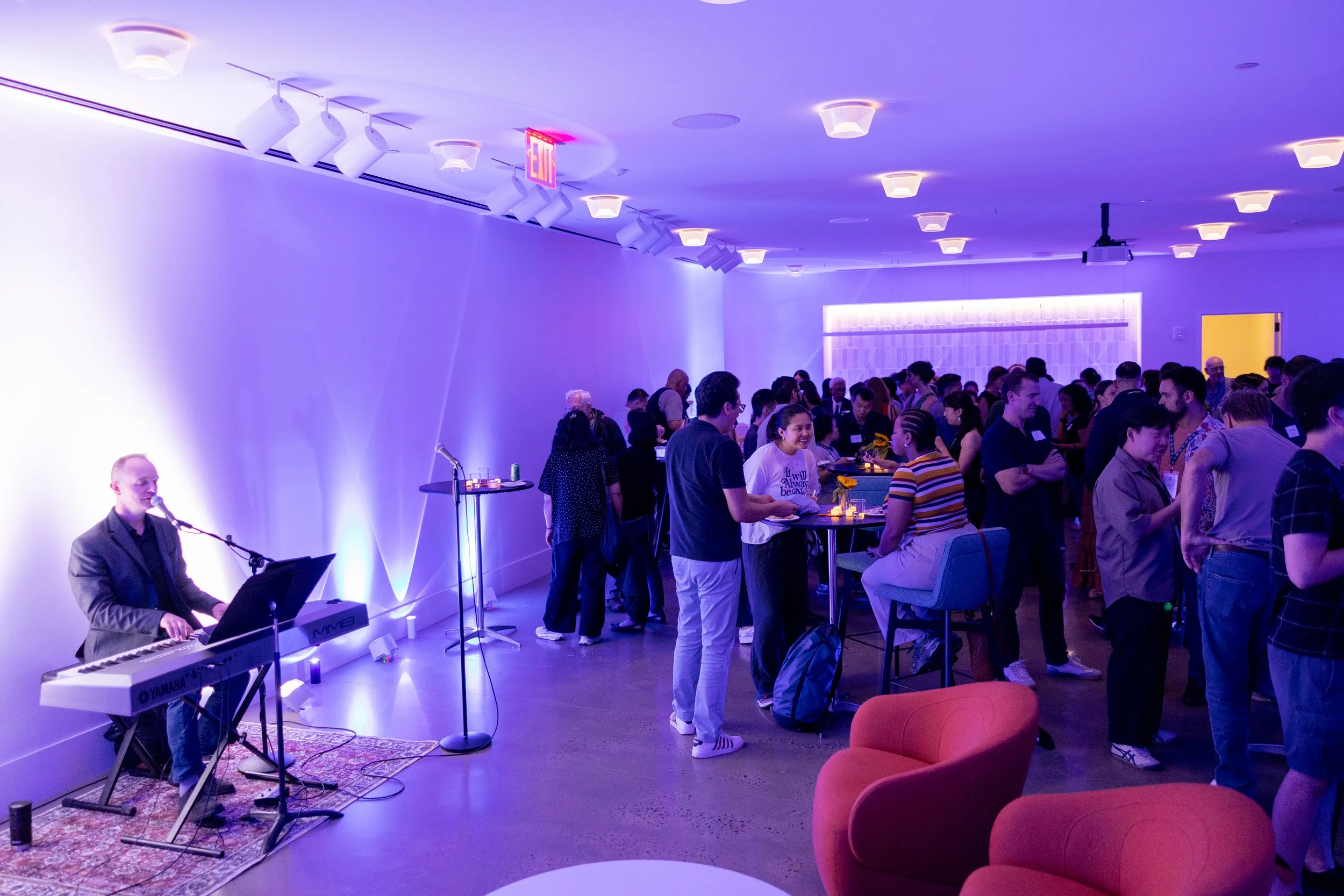 A musical performance at a social gathering or event with people mingling, standing, and sitting in a room illuminated with purple and blue lighting, featuring a keyboard player and a small stage area.