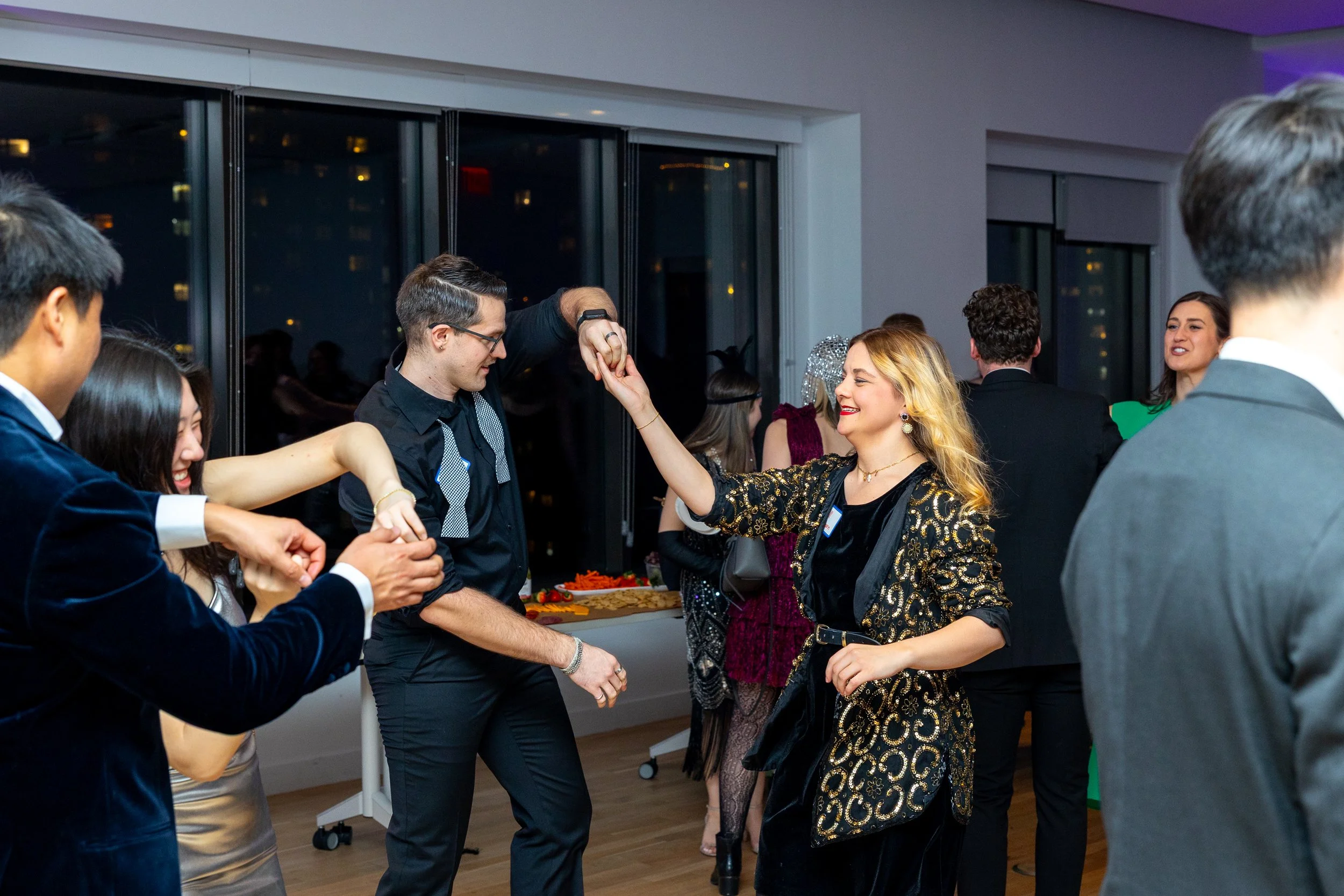 People dancing and socializing at a festive indoor party, with a woman in a black and gold dress smiling and holding hands with a man, while others in the background are enjoying the event.