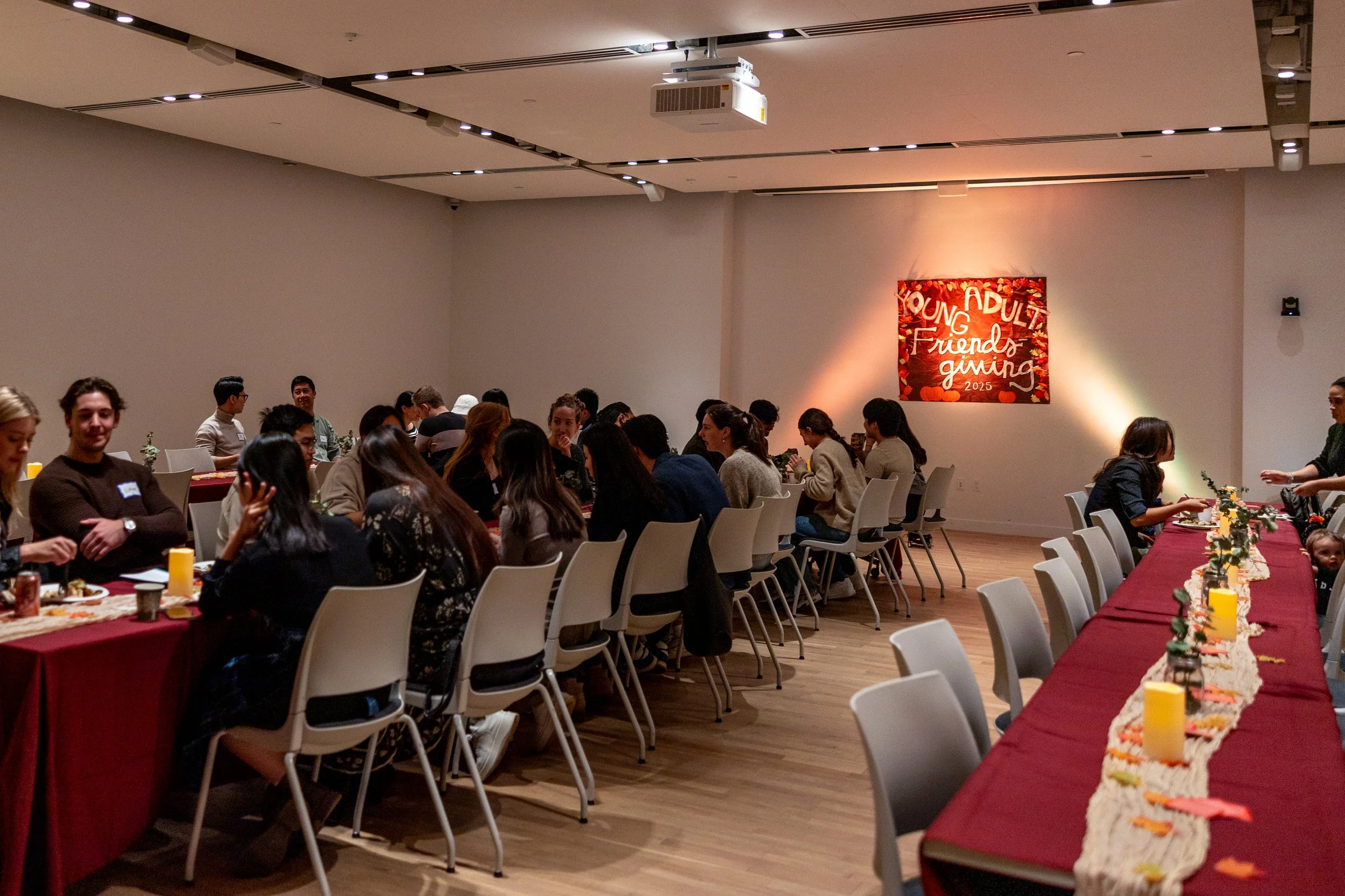 People seated at long dining tables with red tablecloths, decorated with candles and fall-themed decorations, in a banquet hall with a large red and orange sign on the wall.