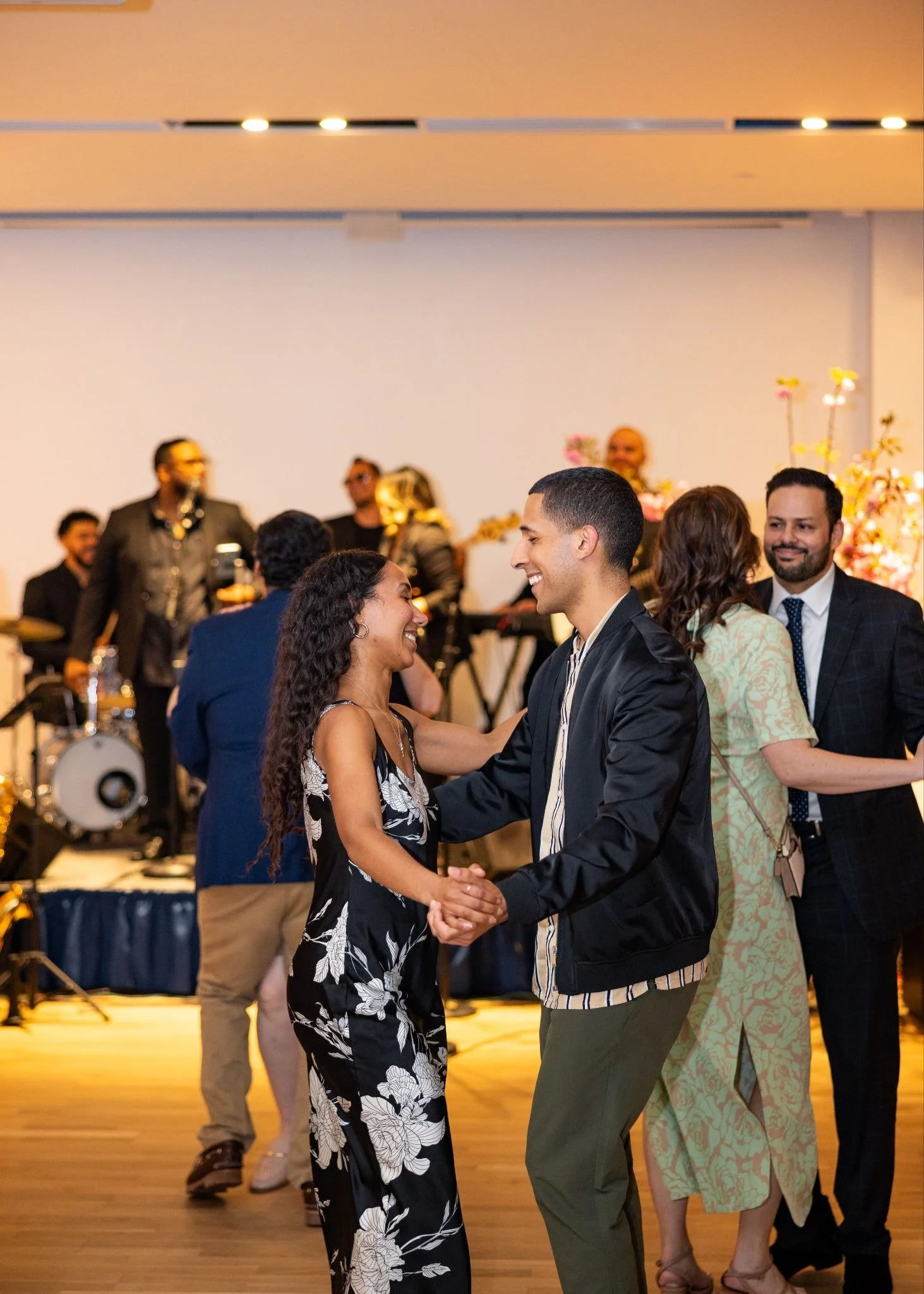 Couple dancing and smiling at a lively indoor celebration or party, with a live band playing in the background and floral decorations.