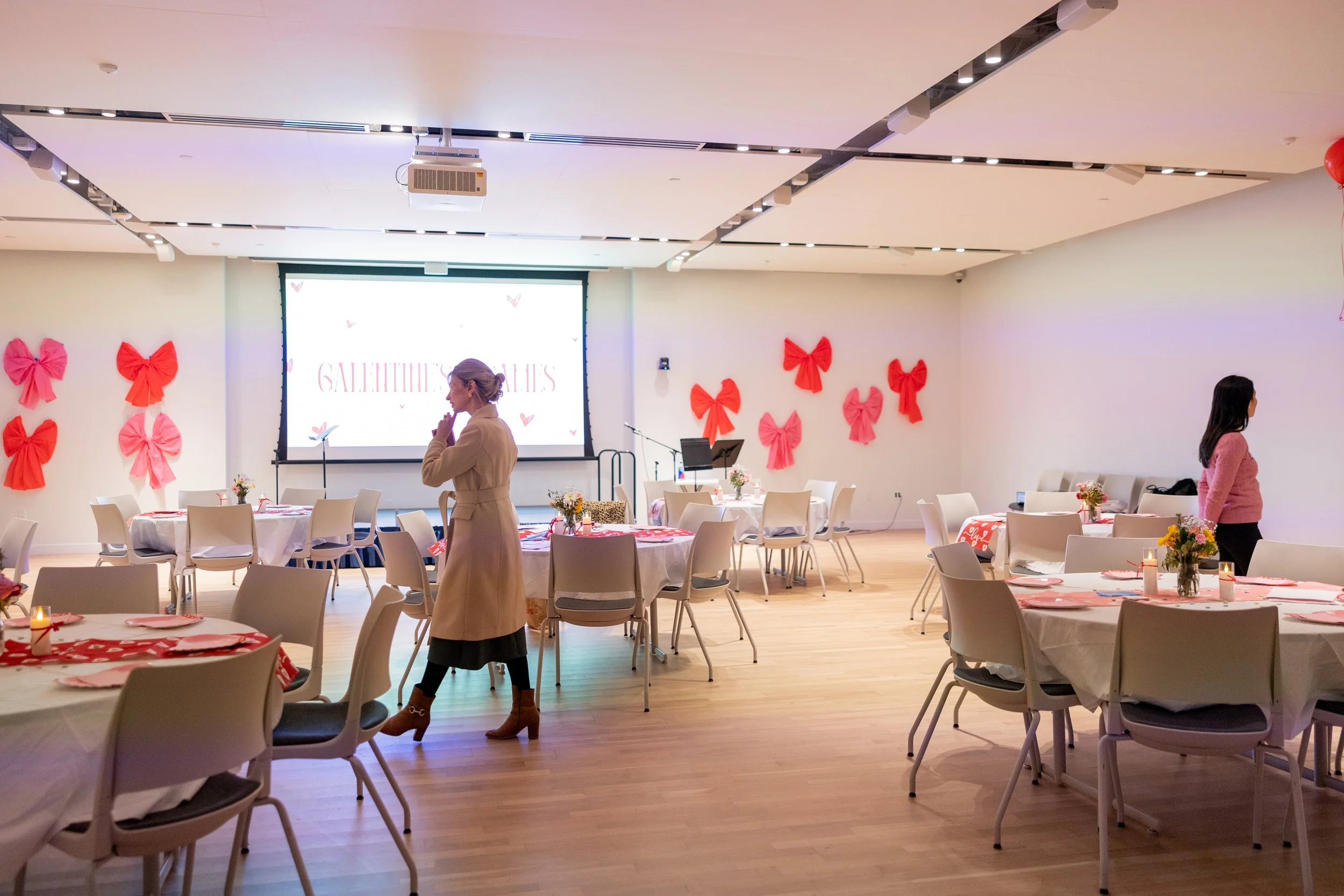A decorated event room with pink and red bows on the walls, set tables with pink napkins and centerpieces, and a large screen at the front. Two women are standing in the room, one near the front and one towards the middle.