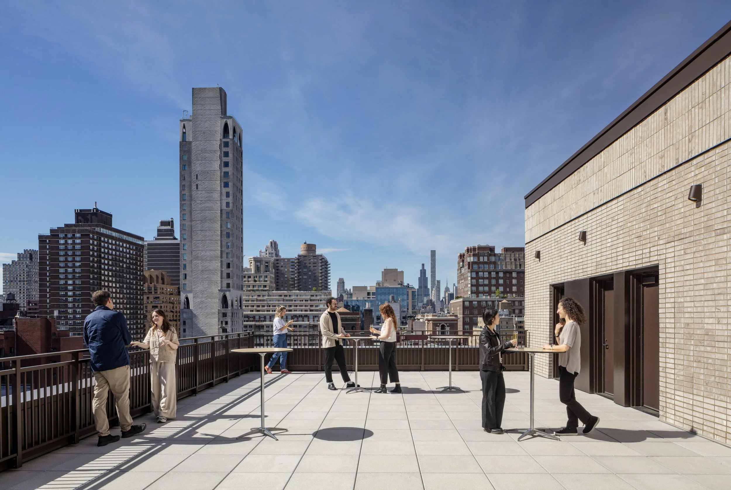 People on a rooftop terrace in a city, engaging in conversations and using phones, with a skyline of tall buildings and a clear blue sky in the background.