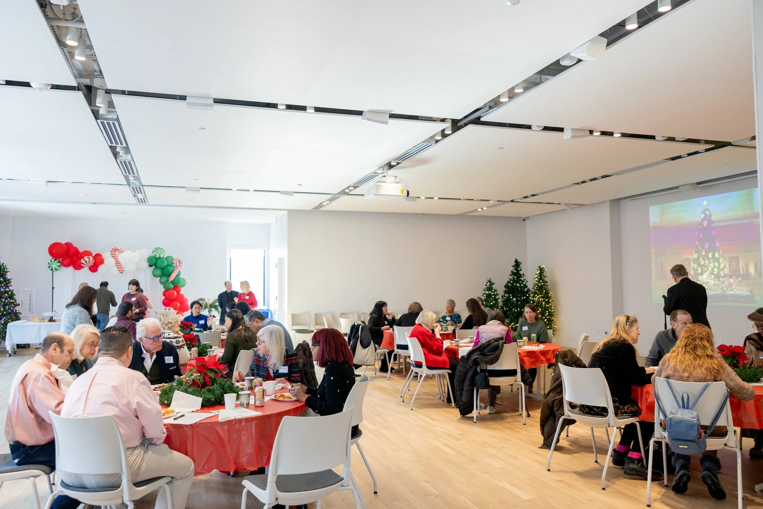 A holiday gathering in a decorated room with Christmas trees, balloon arch, and festive table settings, featuring people seated at round tables, some chatting, enjoying food and drinks, with a large Christmas tree projection on the wall.