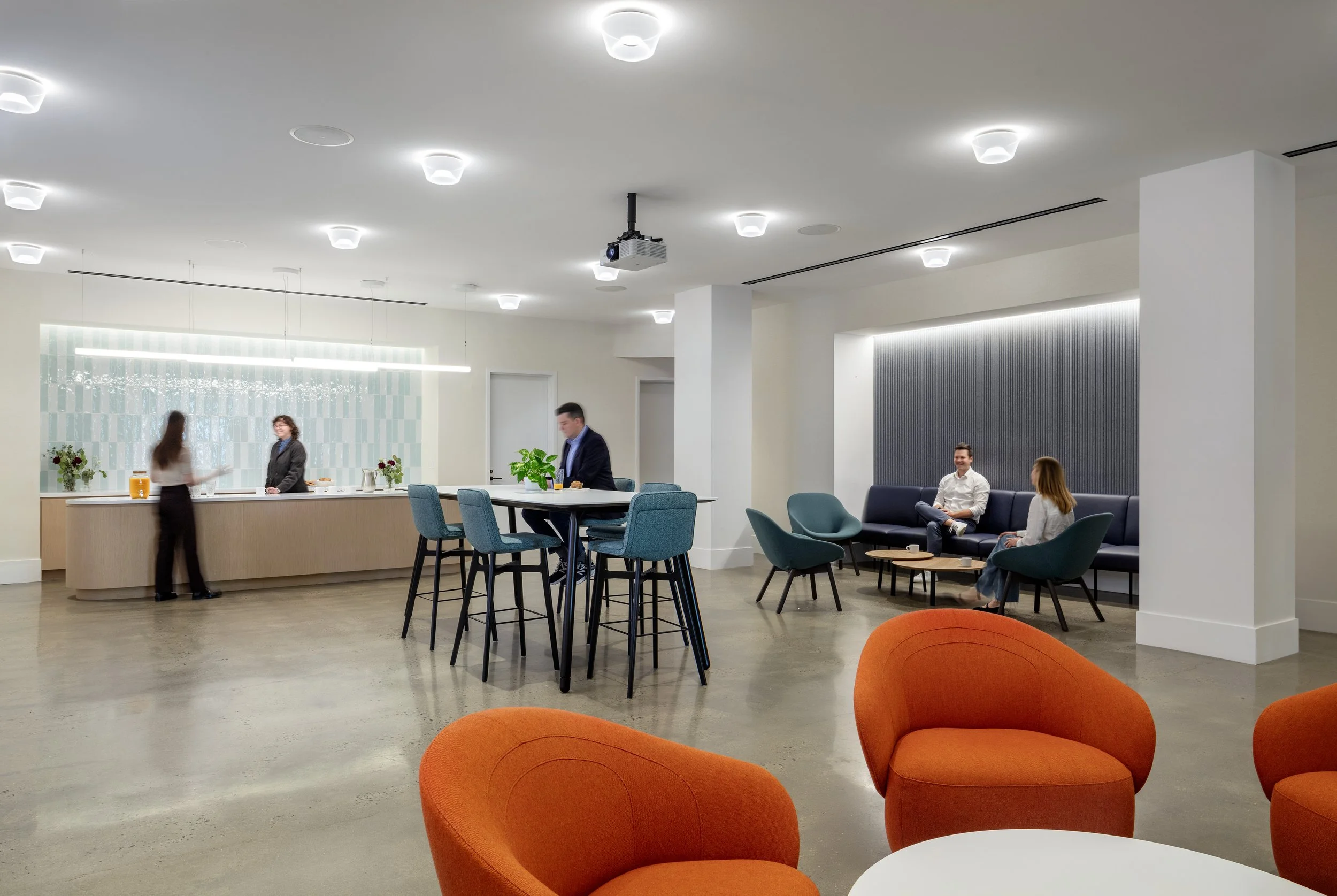 Modern office lounge area with orange chairs in foreground, a high-top table with blue chairs, and a reception counter with staff and a guest, seating area with sofa and armchairs in background.