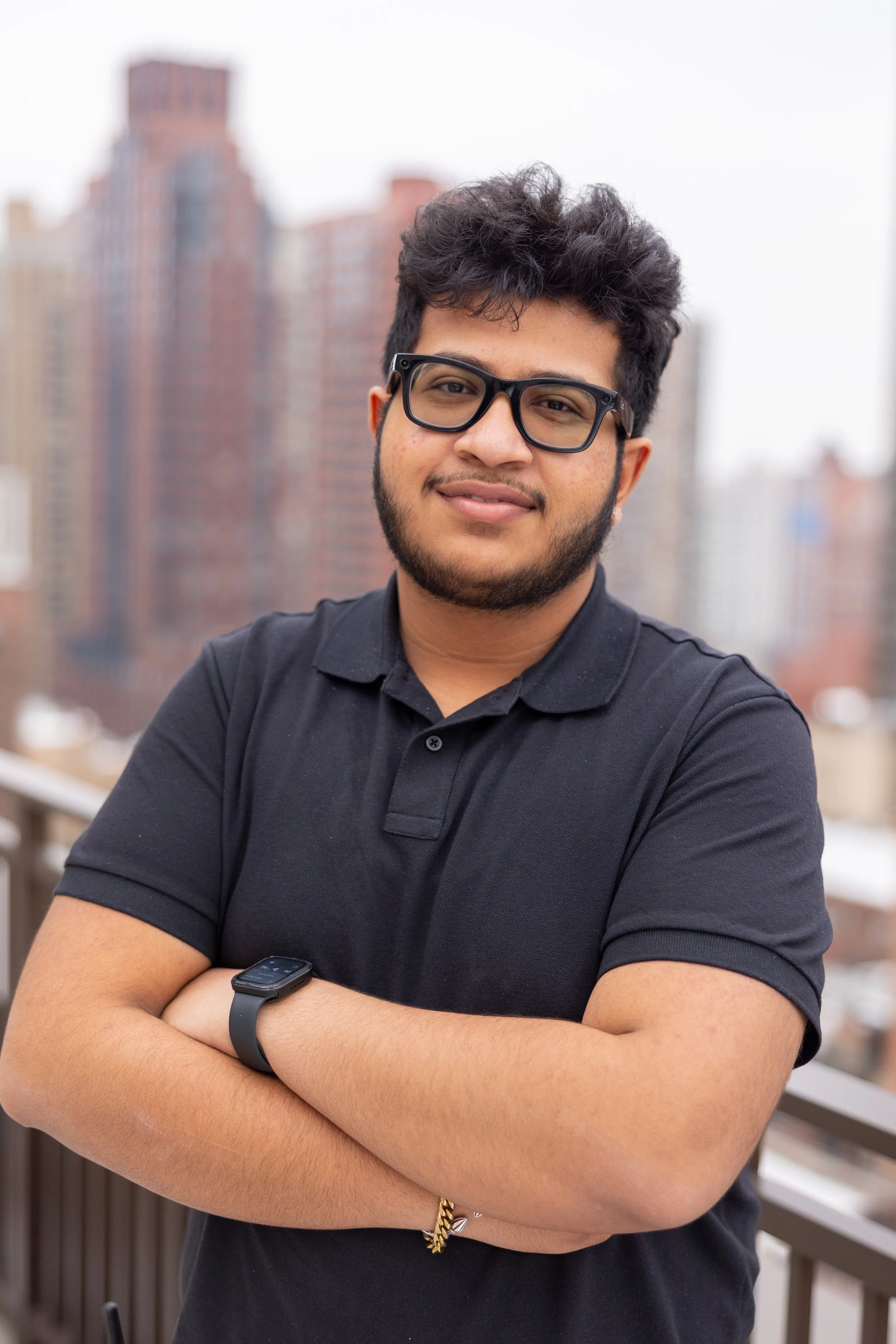 Portrait of a young man with dark curly hair, glasses, and a beard, standing with arms crossed on a balcony with city buildings in the background.
