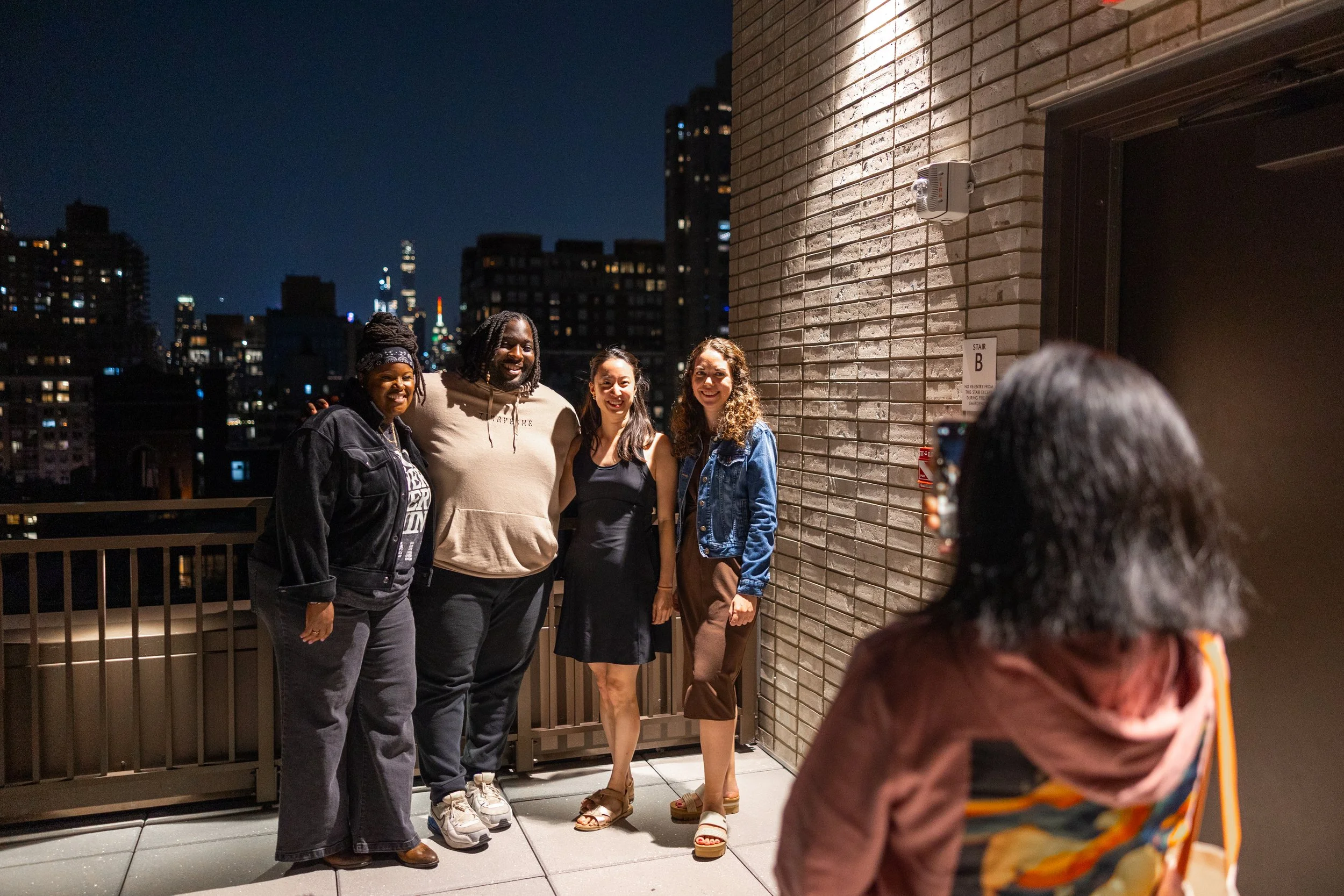 A group of four friends, standing on a balcony at night, shoulder to shoulder, smiling for a photo. A woman with long hair is taking their picture, with her back to the camera. The city skyline is visible in the background.