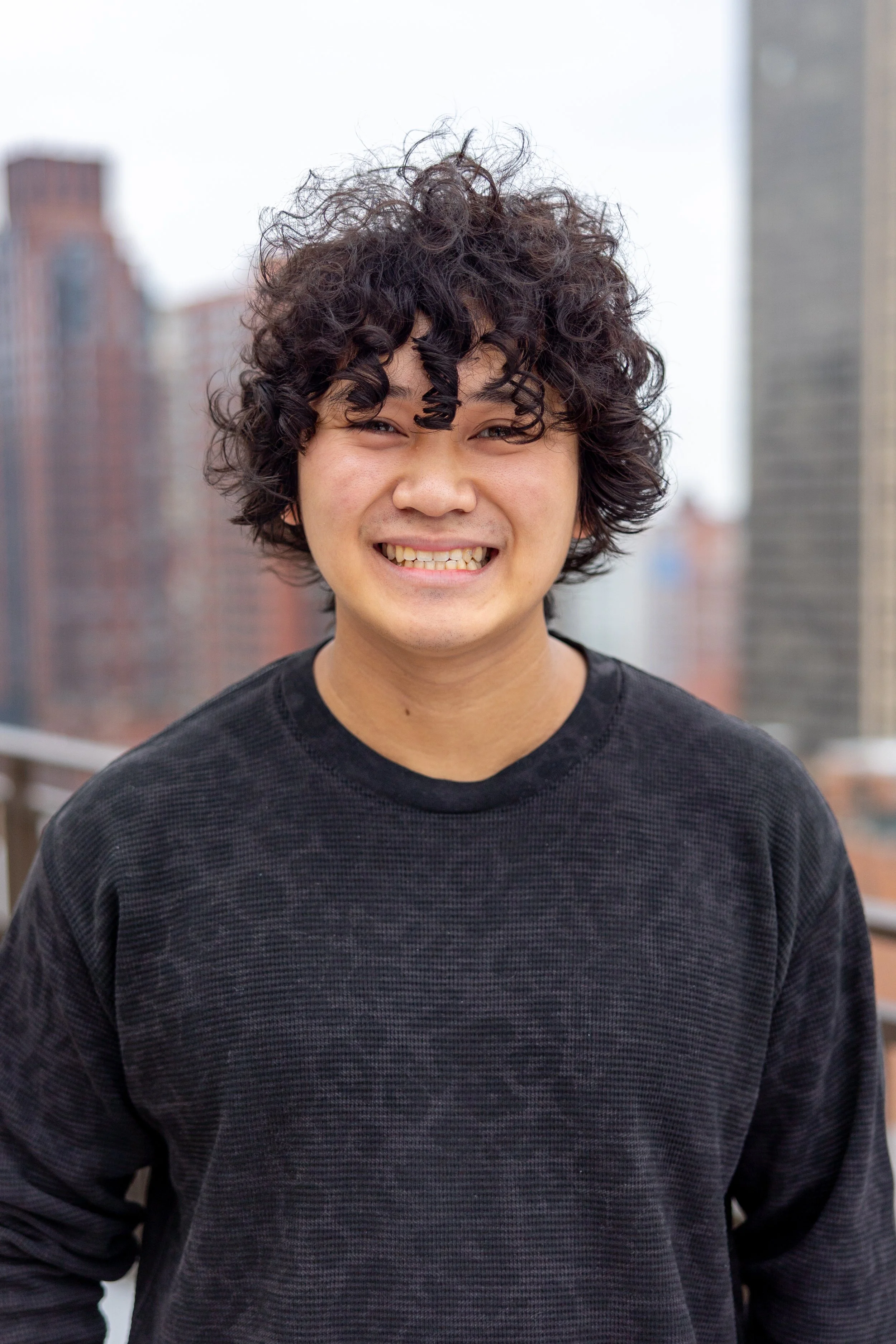 Smiling young man with curly dark hair in a black sweatshirt outdoors with city buildings in the background.