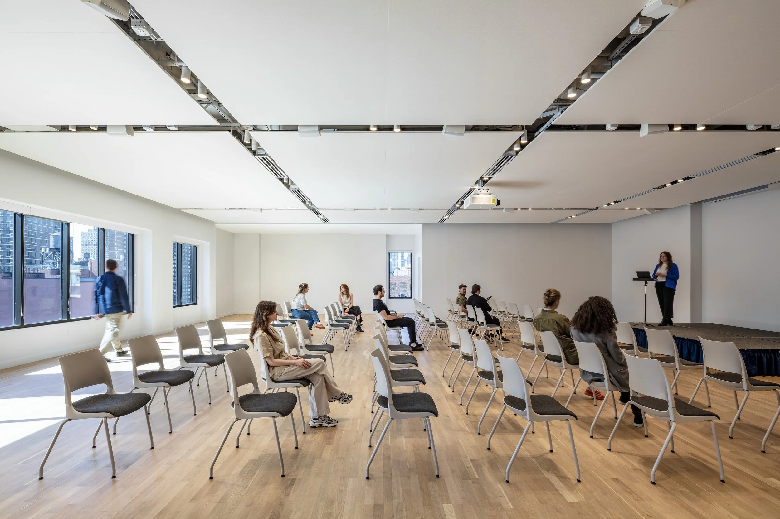 A conference room with chairs arranged in rows and windows letting in natural light. A woman stands at a podium on a stage, preparing to speak to the attendees.