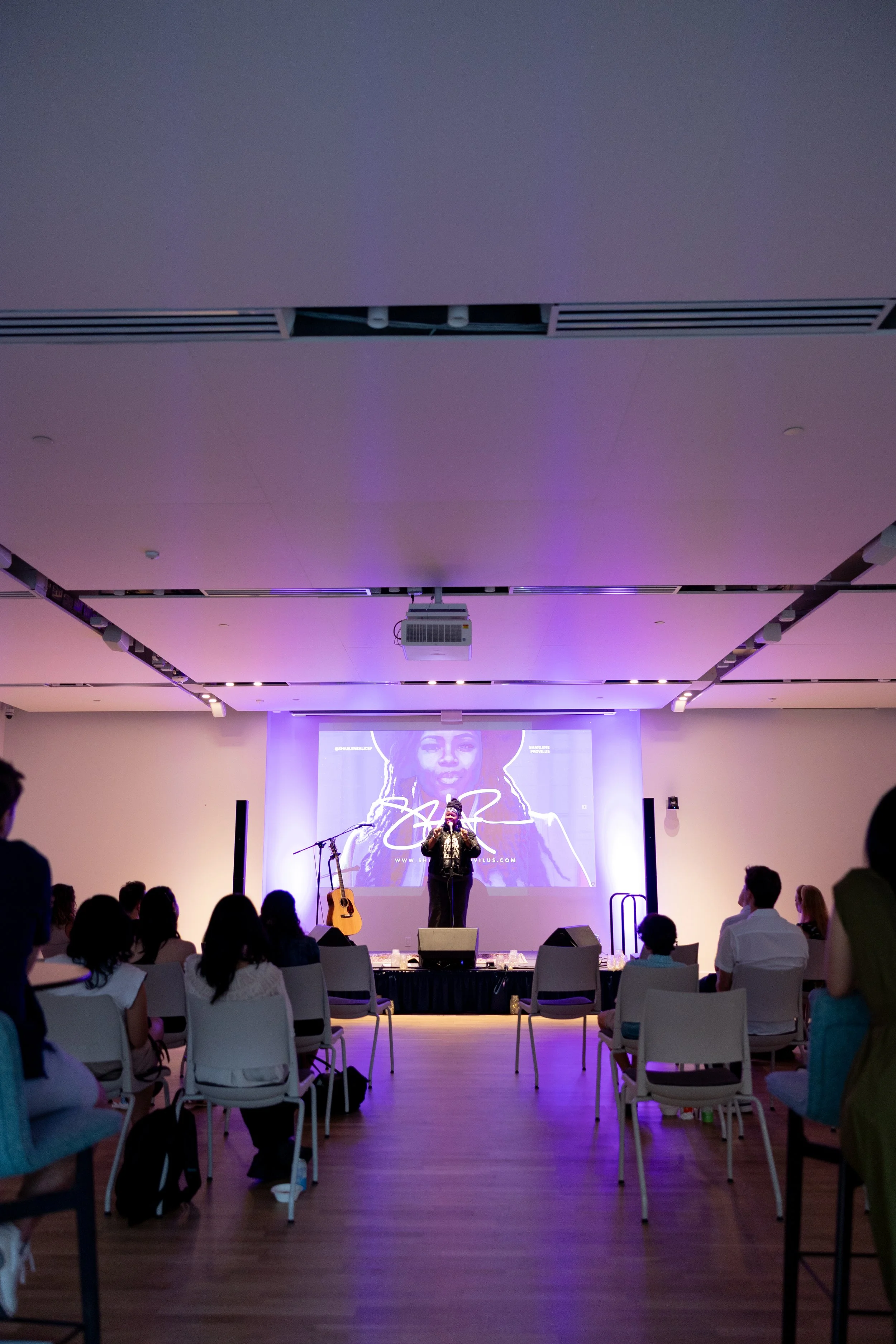 Performer on stage with microphone, guitar on stand, and large screen behind displaying an image of a woman with dreadlocks, in a dimly lit room with audience seated facing the stage.