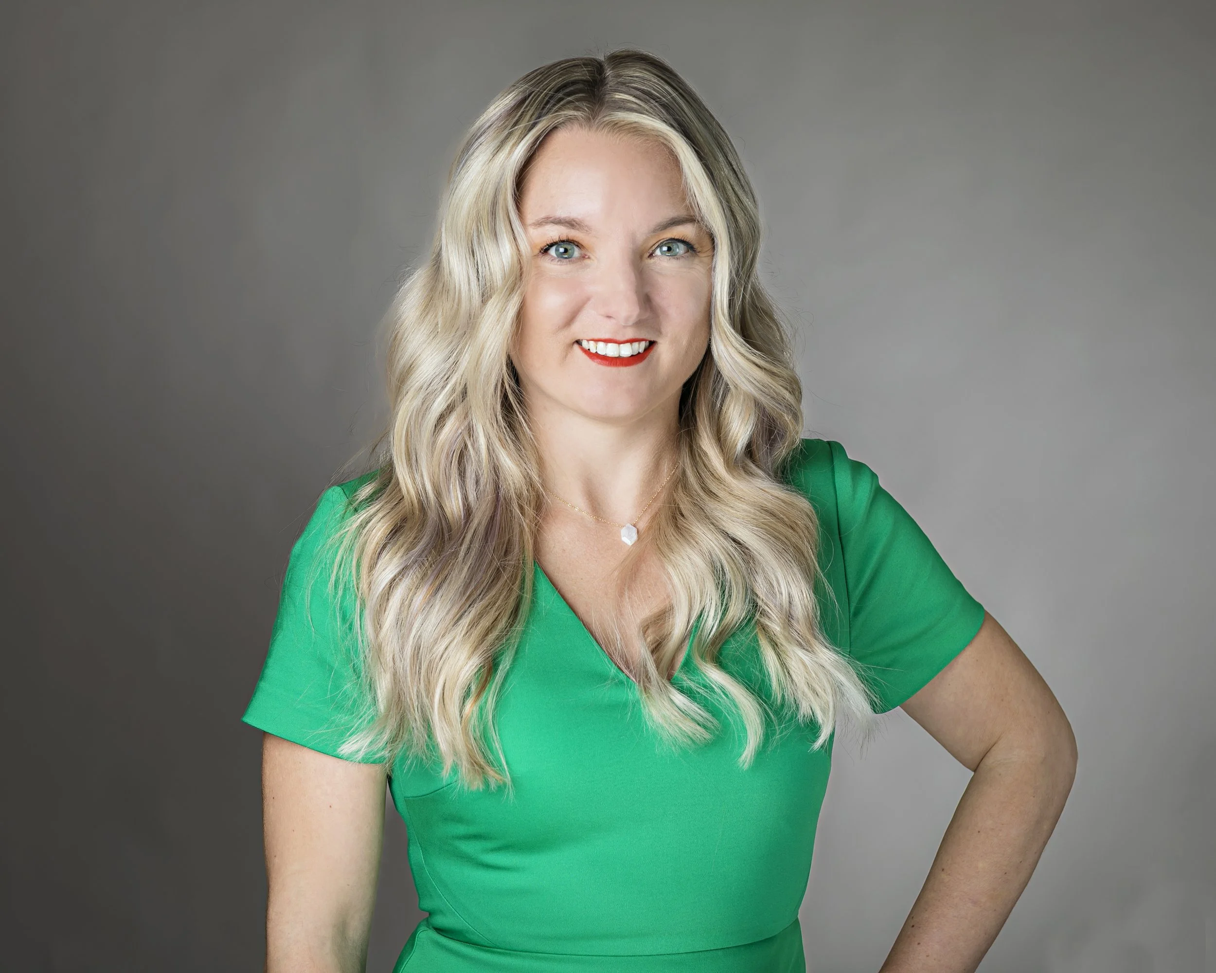 A woman with long, wavy blonde hair wearing a bright green dress, smiling with red lipstick, posed against a neutral gray background.