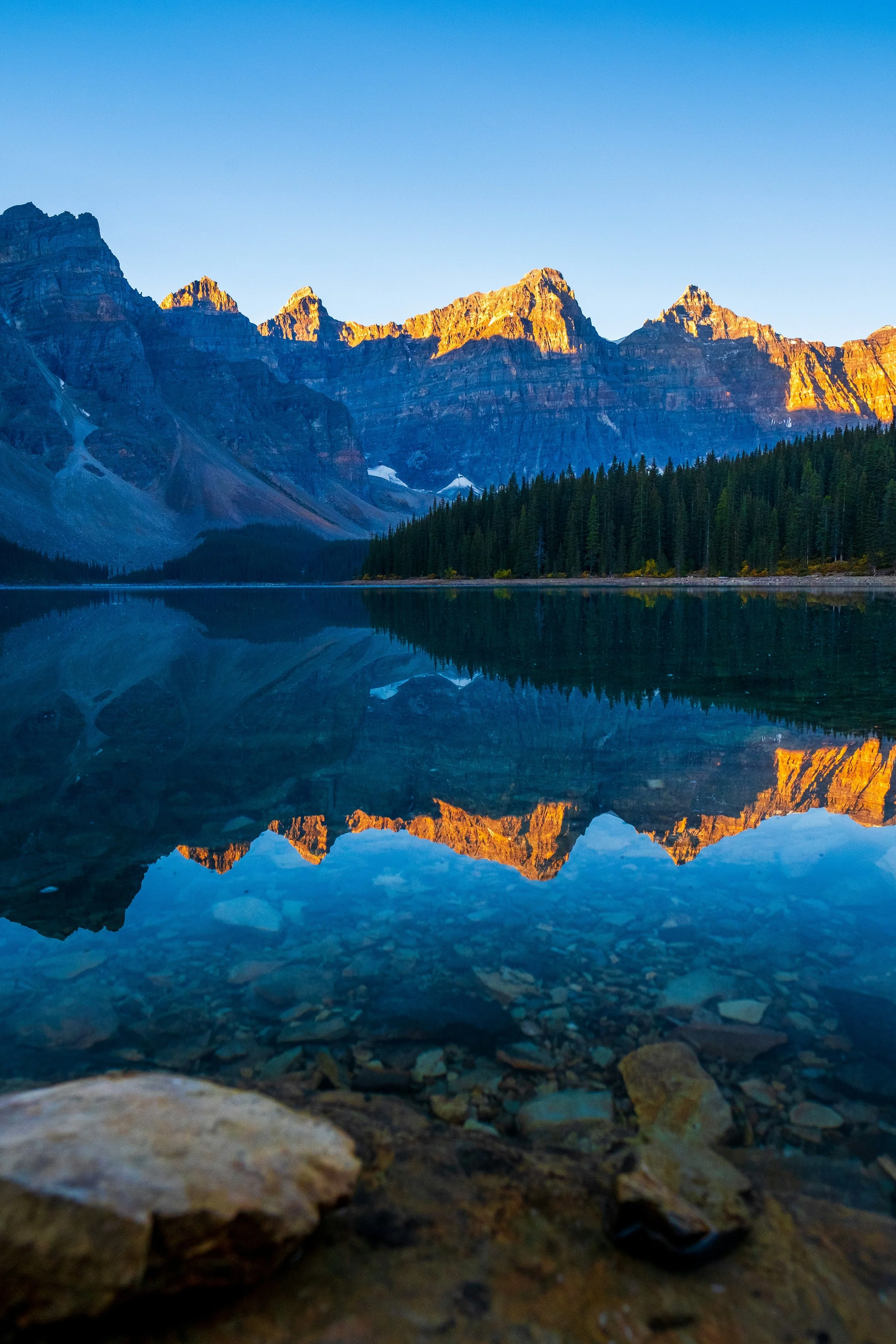 Scenic landscape of a mountain range reflected in a calm lake at sunrise or sunset, with trees along the shoreline.