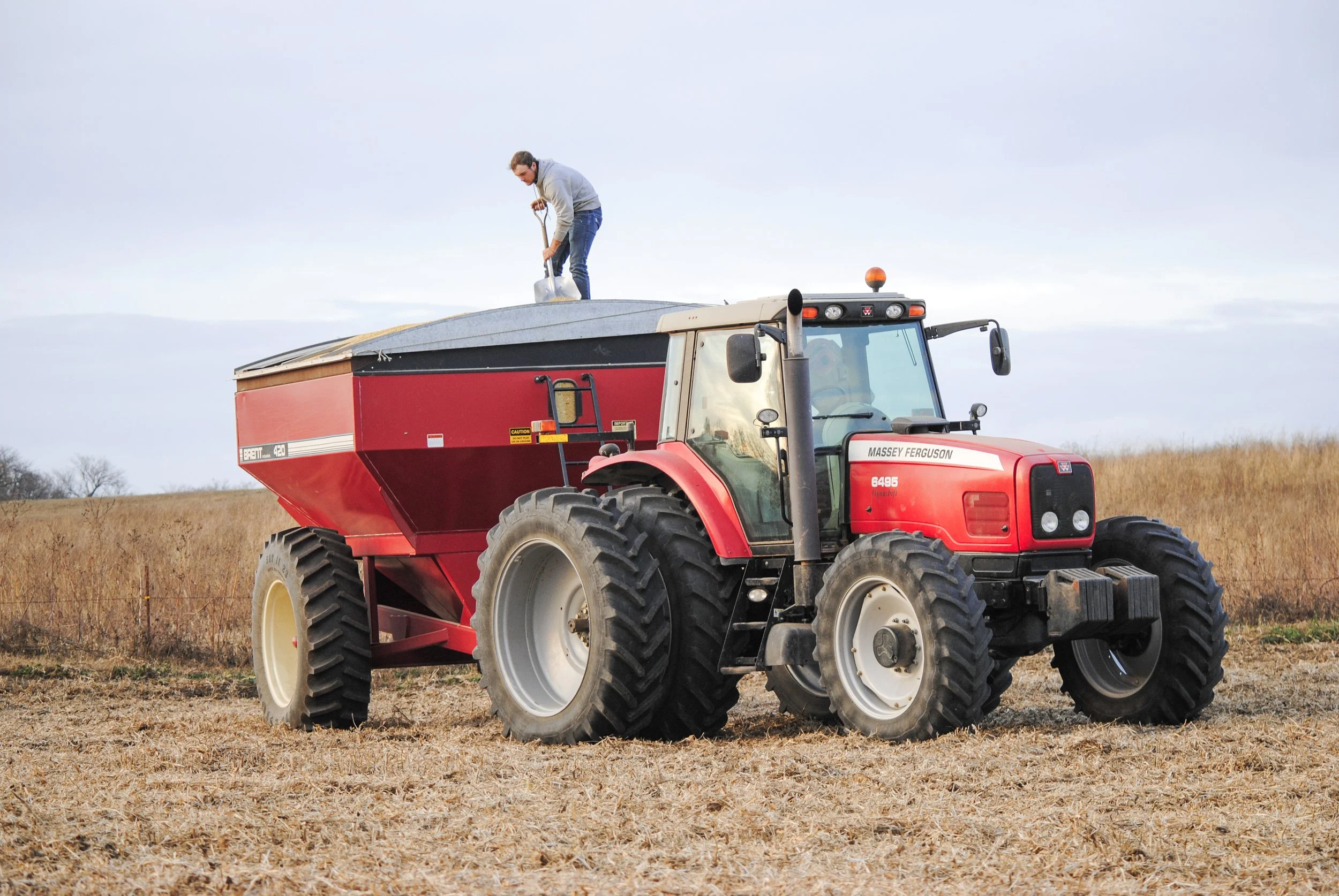 A man standing on top of a large red tractor, filling a component with a tool in a field with dry grass and a partly cloudy sky.