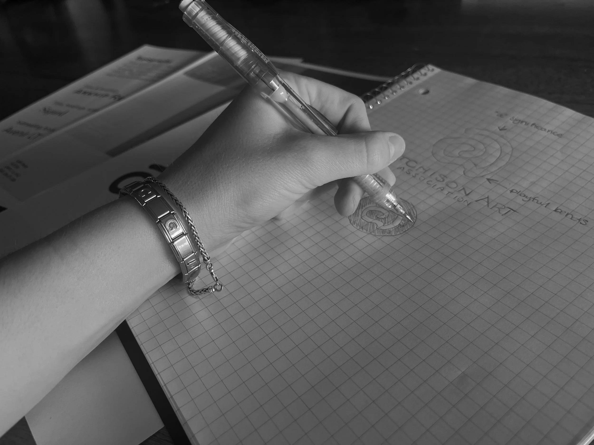 A person writing in a notebook with a pen, with a printed brochure or flyer beneath their hand and a spiral notebook beside. The person is wearing bracelets on their wrist.