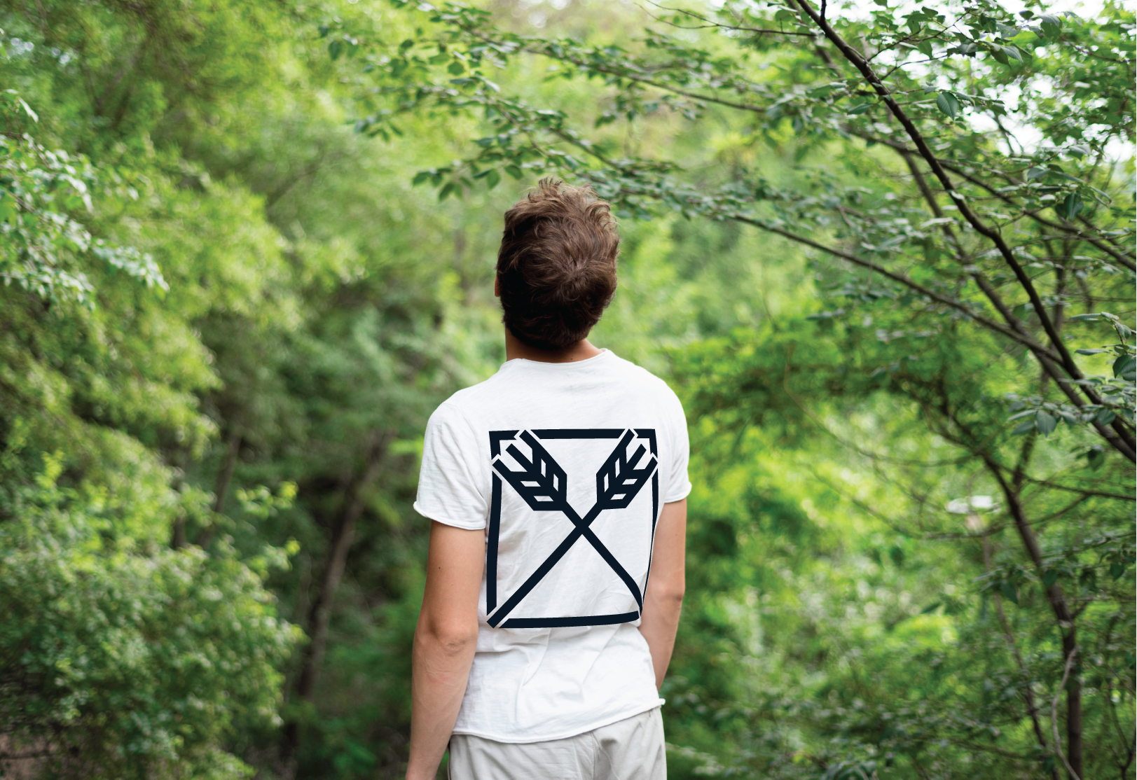 Person with short hair standing in a lush green forest, facing away, wearing a white T-shirt with a black graphic of arrows and leaves on the back.