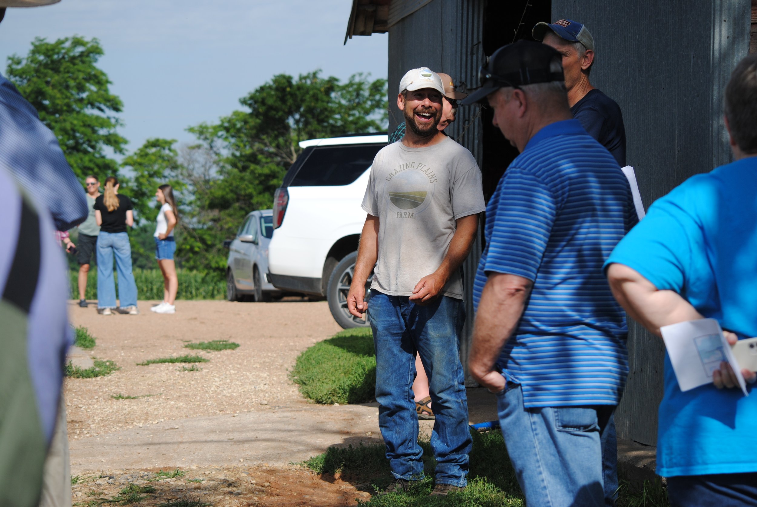 Group of people standing outside near a building, some engaging in conversation, with cars and green trees in the background.