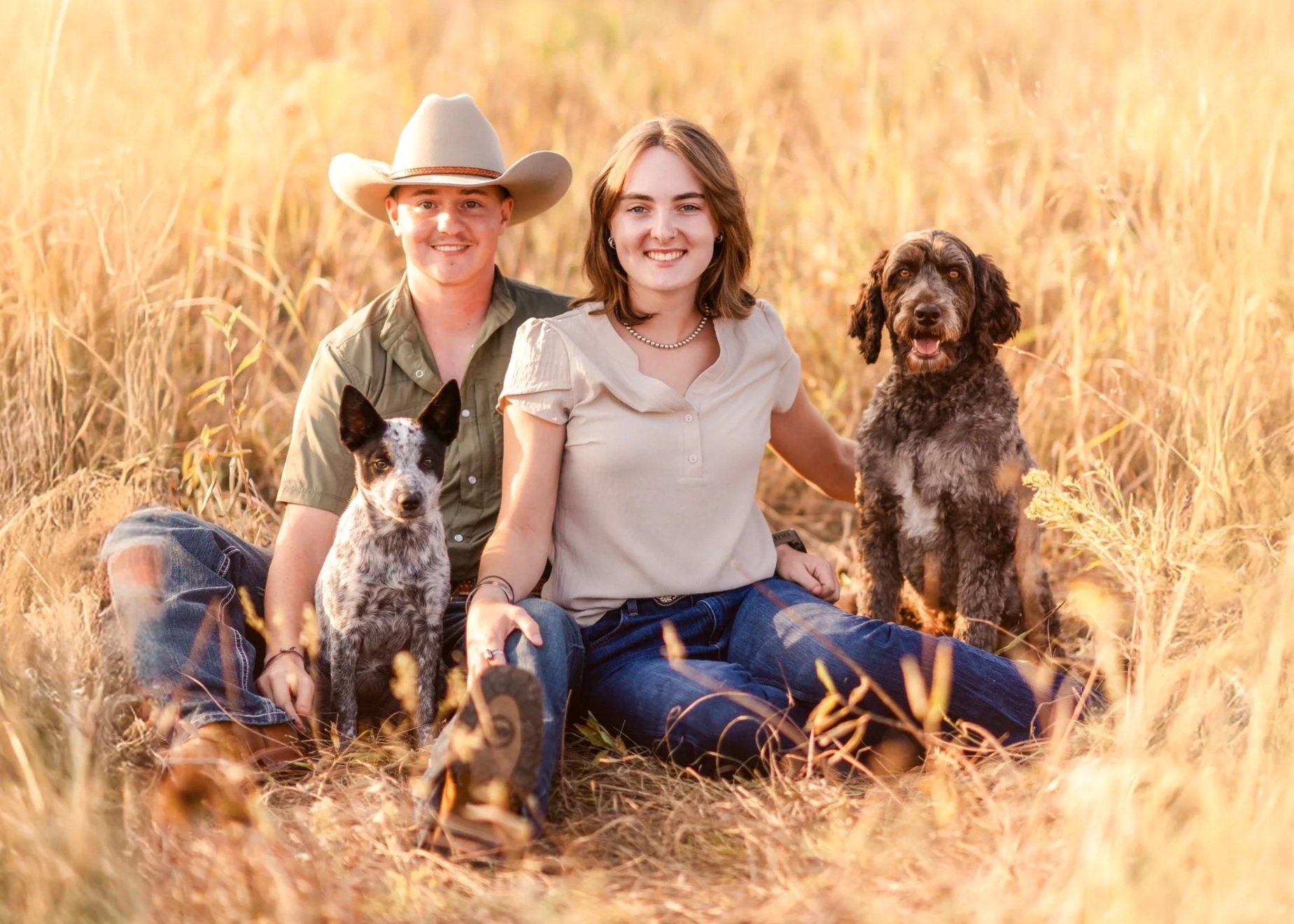 A young man and woman sitting in a field with two dogs during sunset, smiling at the camera.