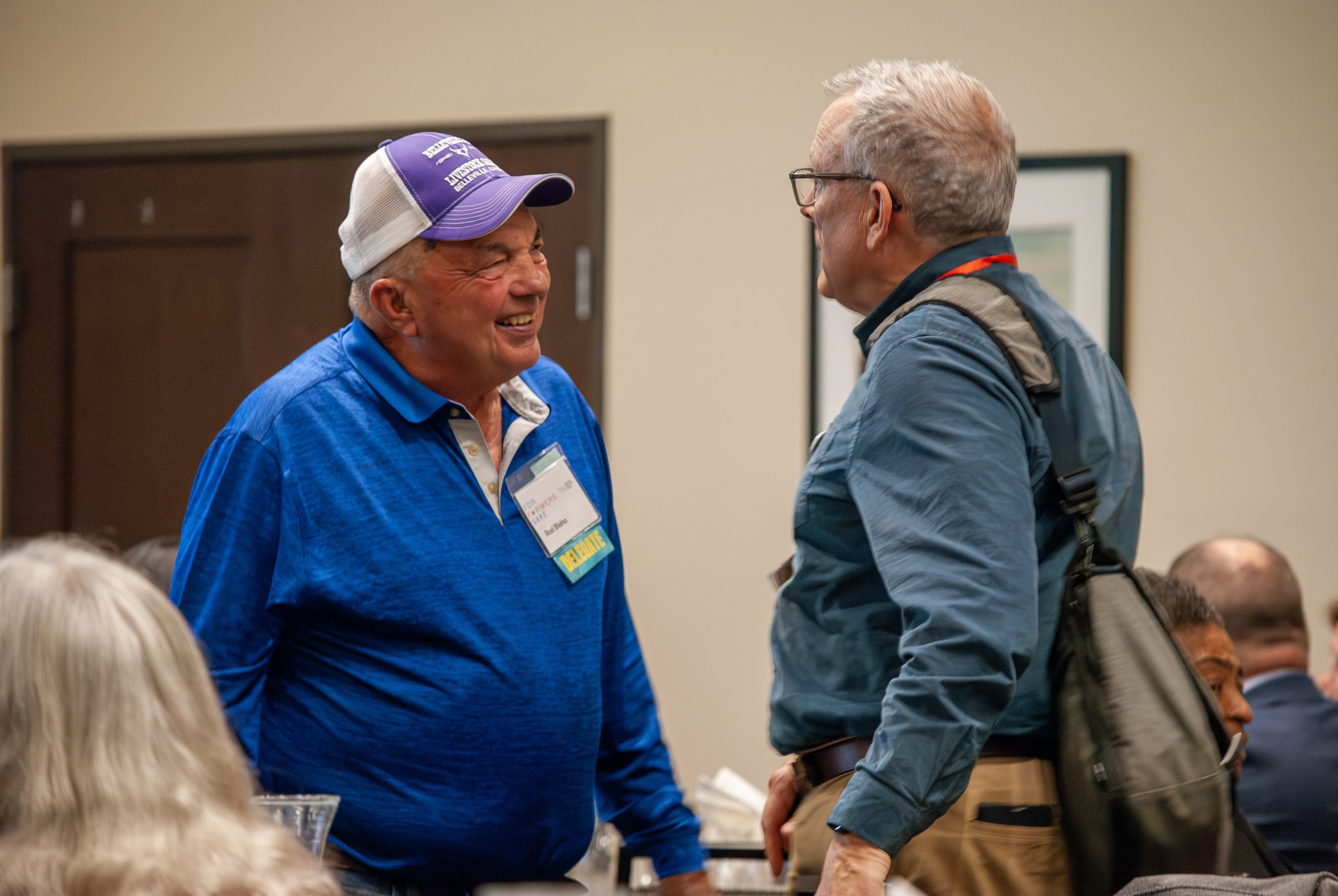 Two men are smiling and talking to each other at a conference or gathering, with other people seated around them.