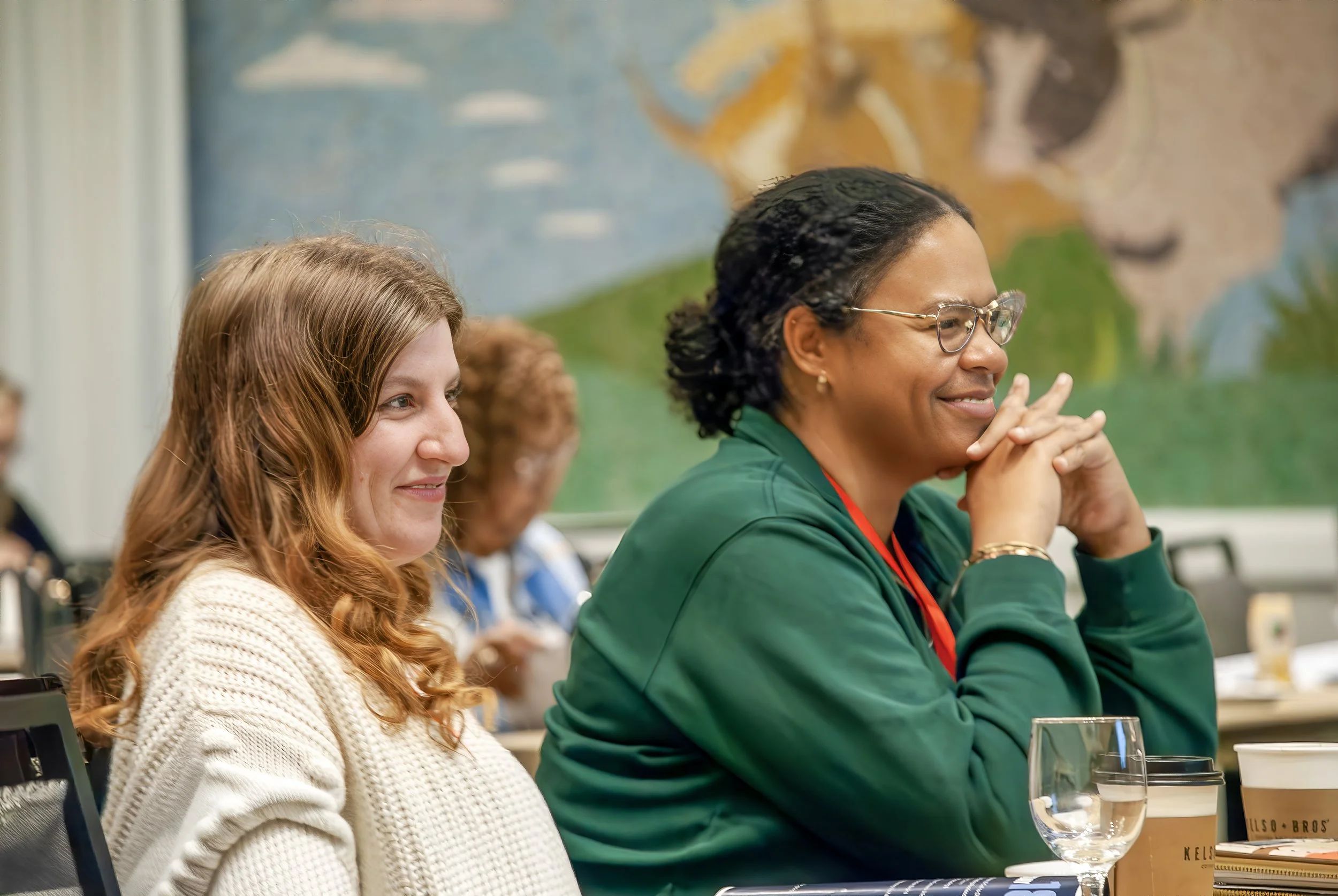 Two women sitting at a table during a meeting, smiling, with a colorful mural in the background.
