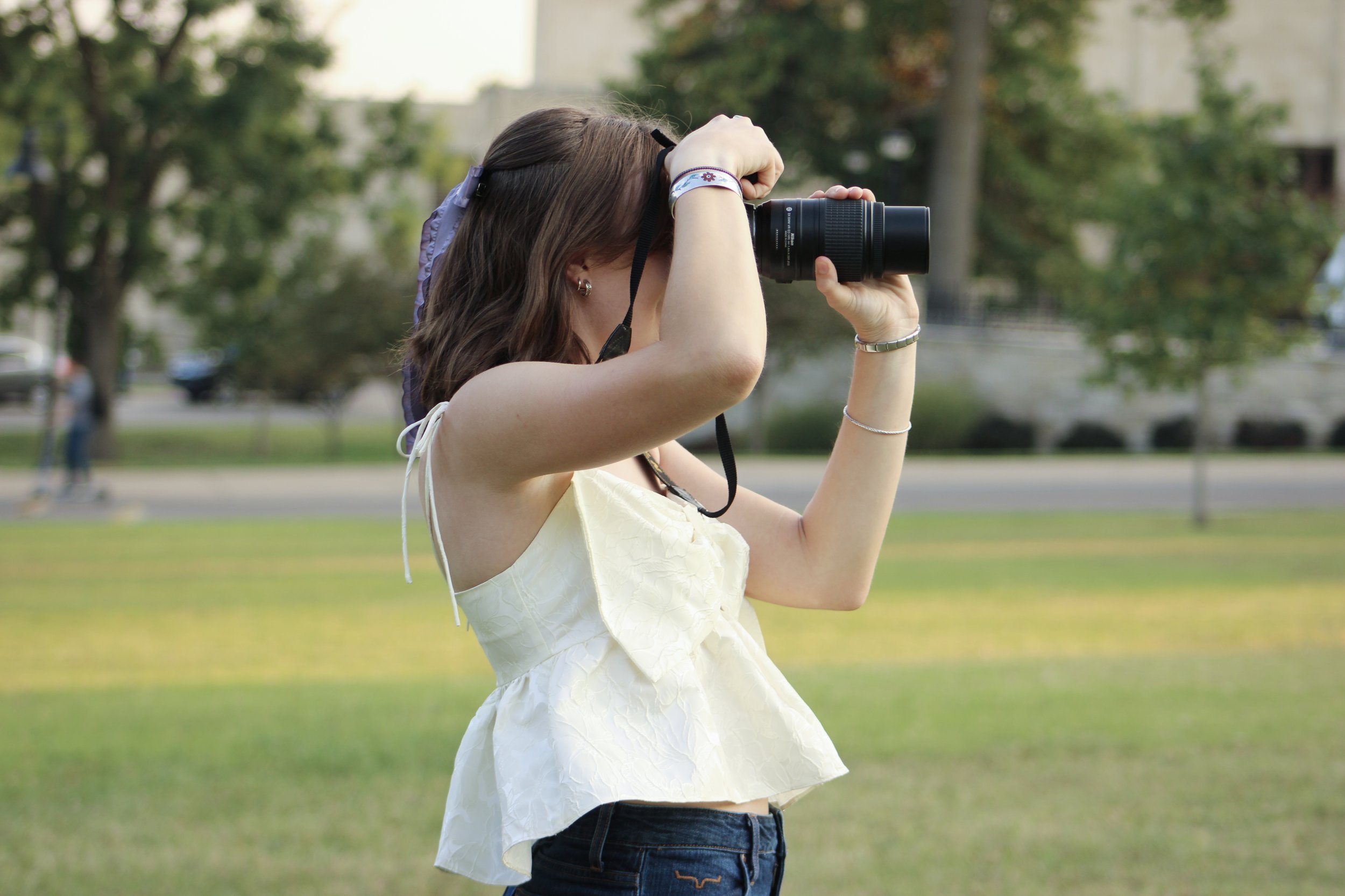 A young woman with brown hair, wearing a cream-colored sleeveless top and dark jeans, is taking a photograph outdoors with a camera in a park during daytime.
