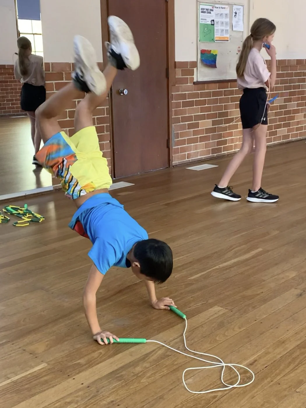 Child performing a handstand on the wooden floor in a gym or classroom, with a jump rope nearby. Other children are standing against the wall.