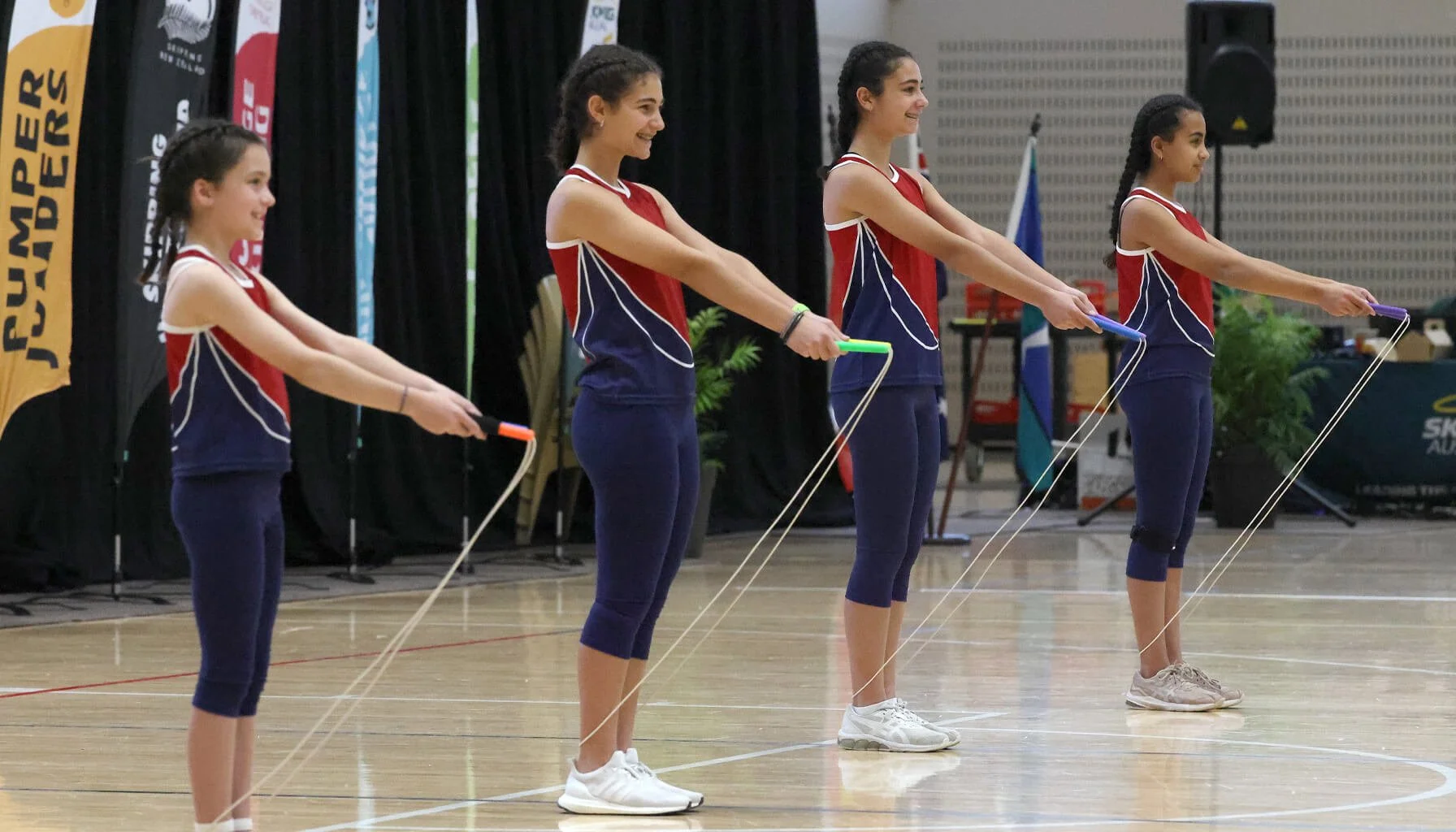 Four young female gymnasts in matching red and blue uniforms practicing with small elastic jump ropes on a gymnasium floor.