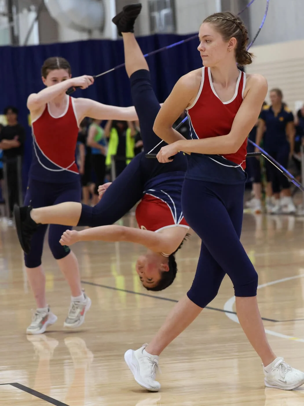 Two young female athletes in red and blue sports uniforms are practicing for a sport drill inside a gymnasium. One girl is holding a broken pole, and the other is swinging the pole, with a third girl falling in front of them.