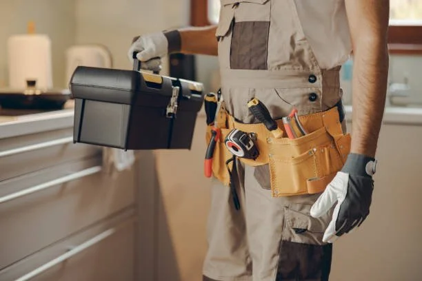 Close-up of a maintenance worker wearing gloves and a tool belt, holding a toolbox in a home setting.