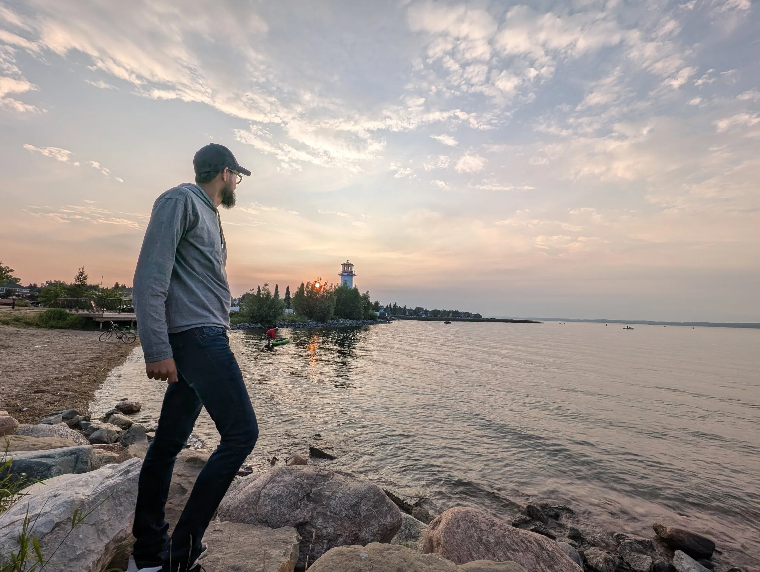 A man in a gray hoodie and black cap walking along a rocky lakeshore during sunset, with a lighthouse and trees in the background.