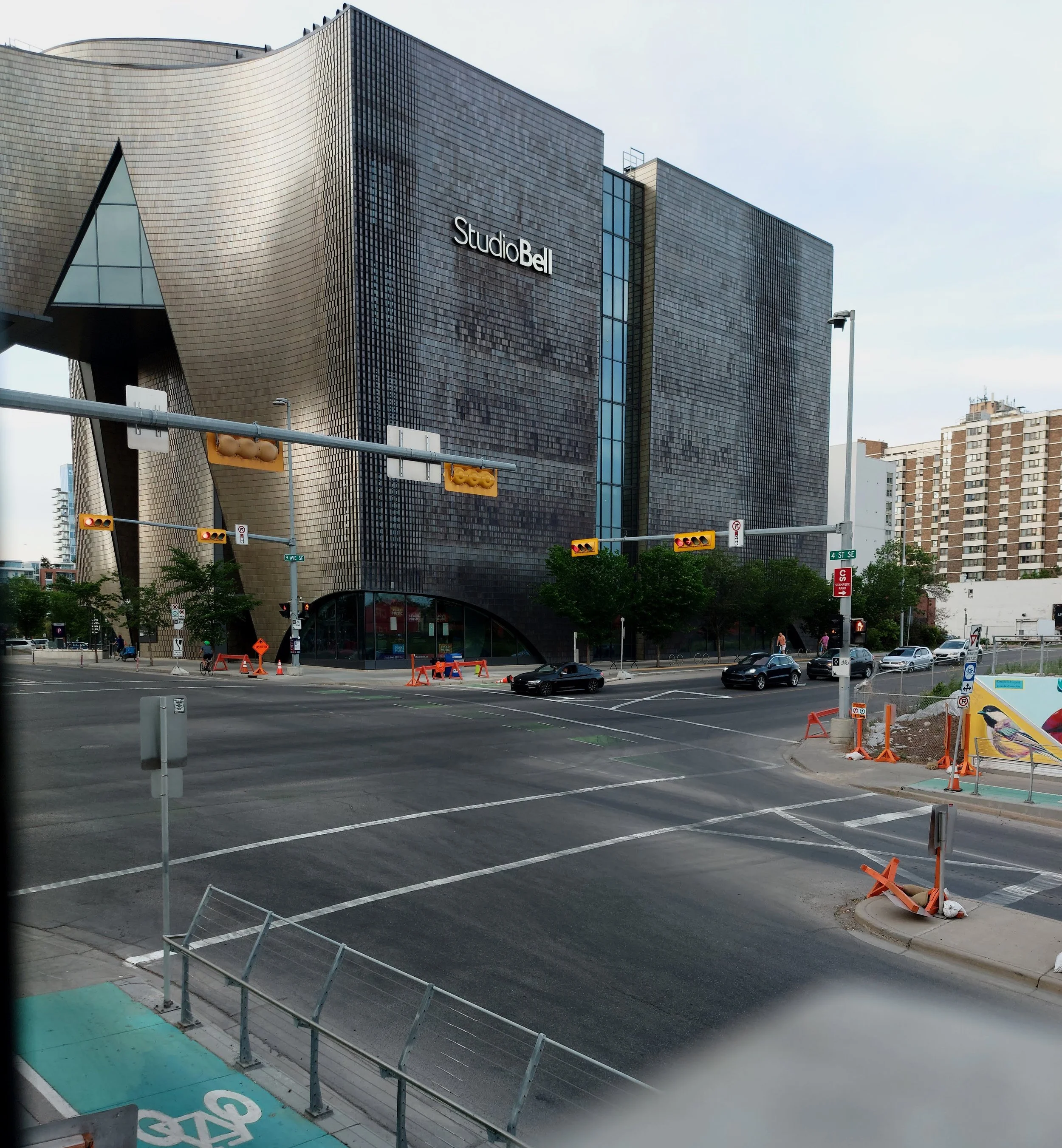 View of StudioBell building with black and gold exterior, city street, cars, and pedestrians, under cloudy sky.