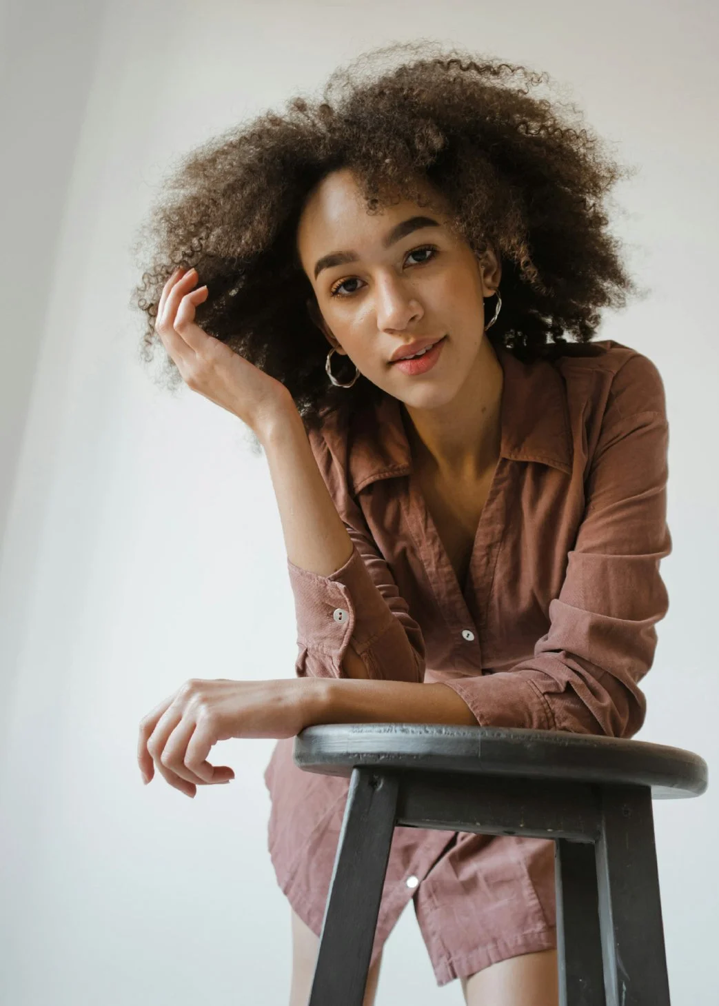 Young woman with curly hair, wearing hoop earrings and a brown button-up shirt, sitting on a black wooden stool and resting her arm on it, looking at the camera with a slight smile.