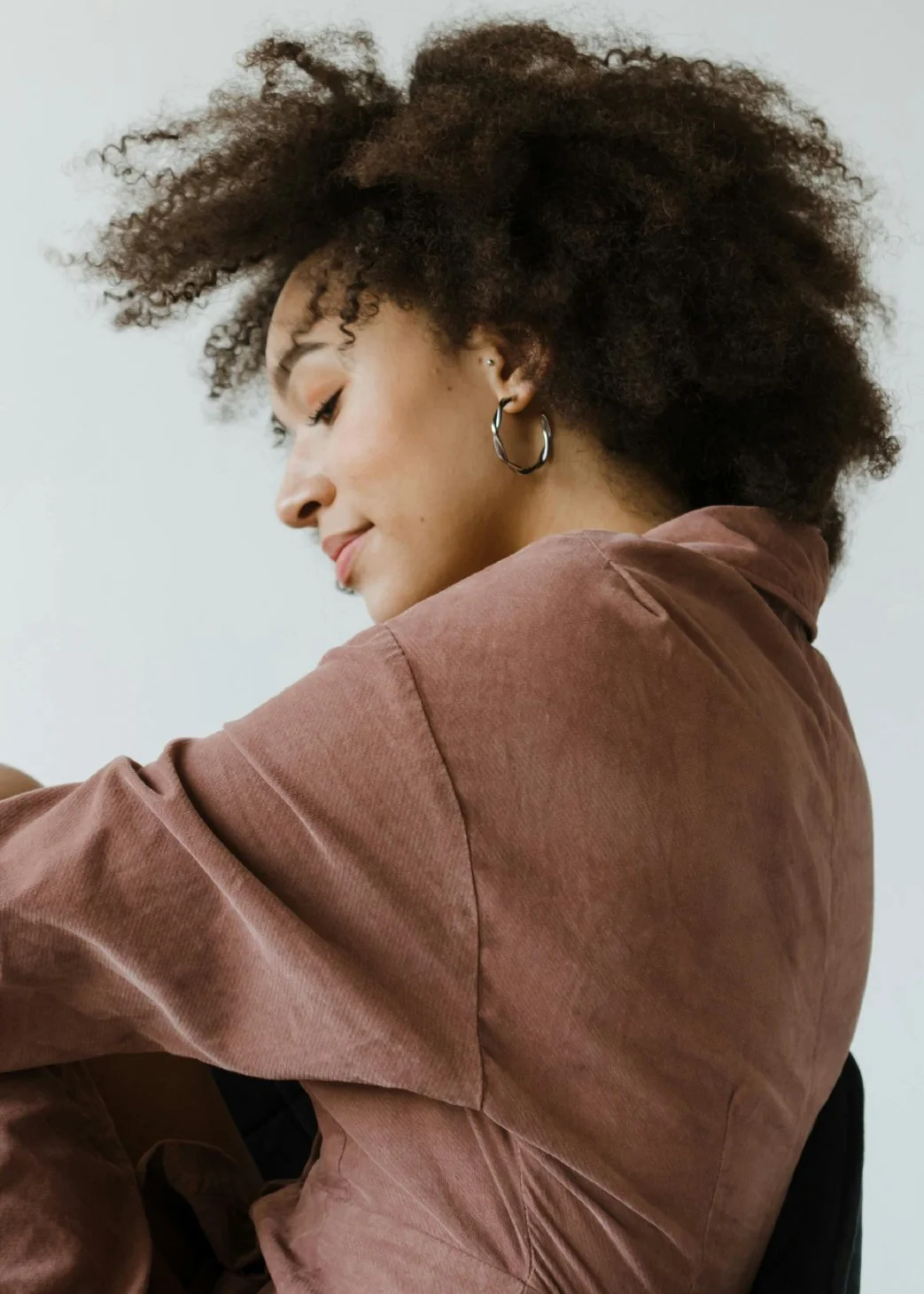A woman with curly hair wearing hoop earrings and a brown shirt, smiling softly with her eyes closed.
