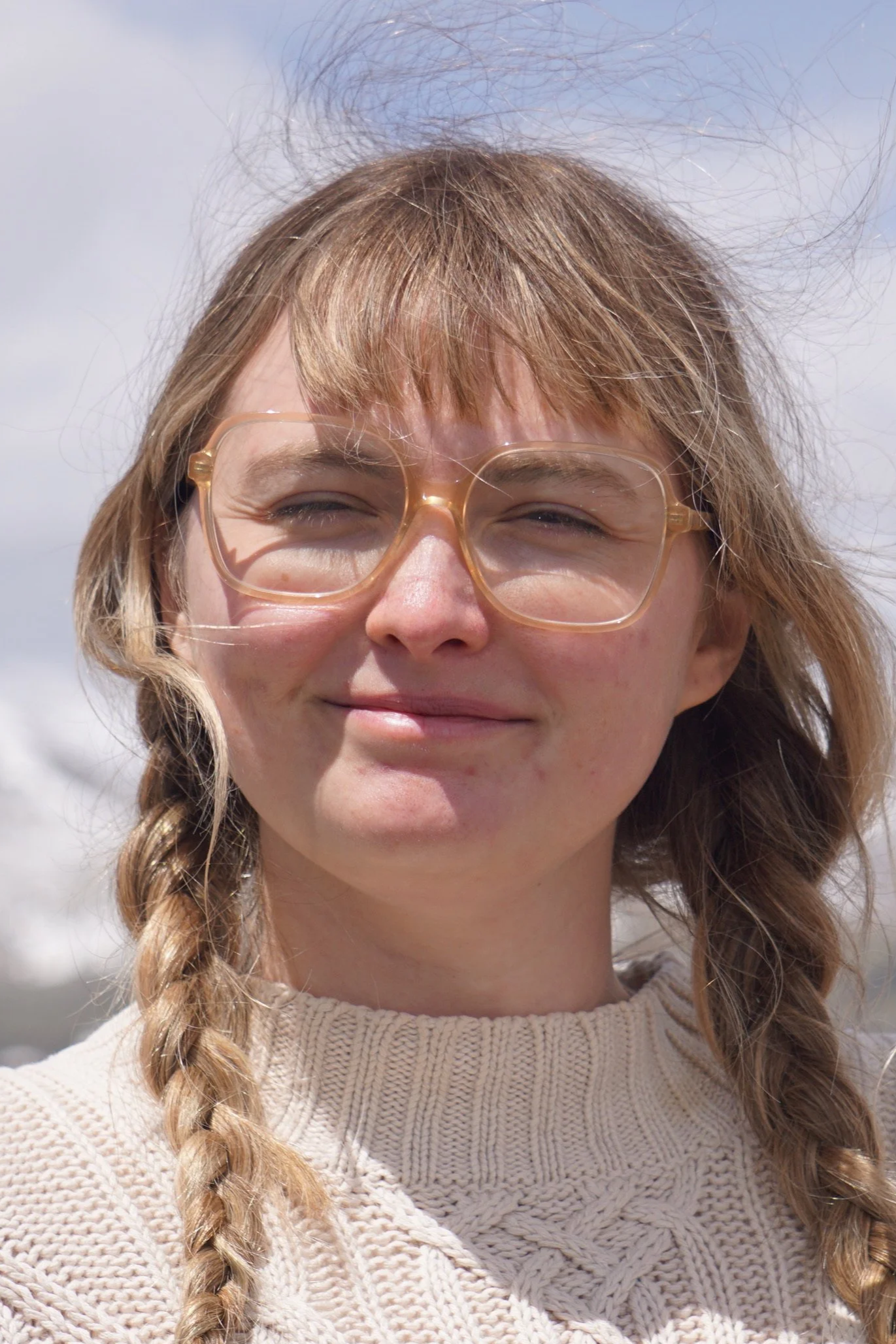 A smiling person with curly hair wearing glasses and a beige knitted sweater, standing outdoors with a bright sky background.