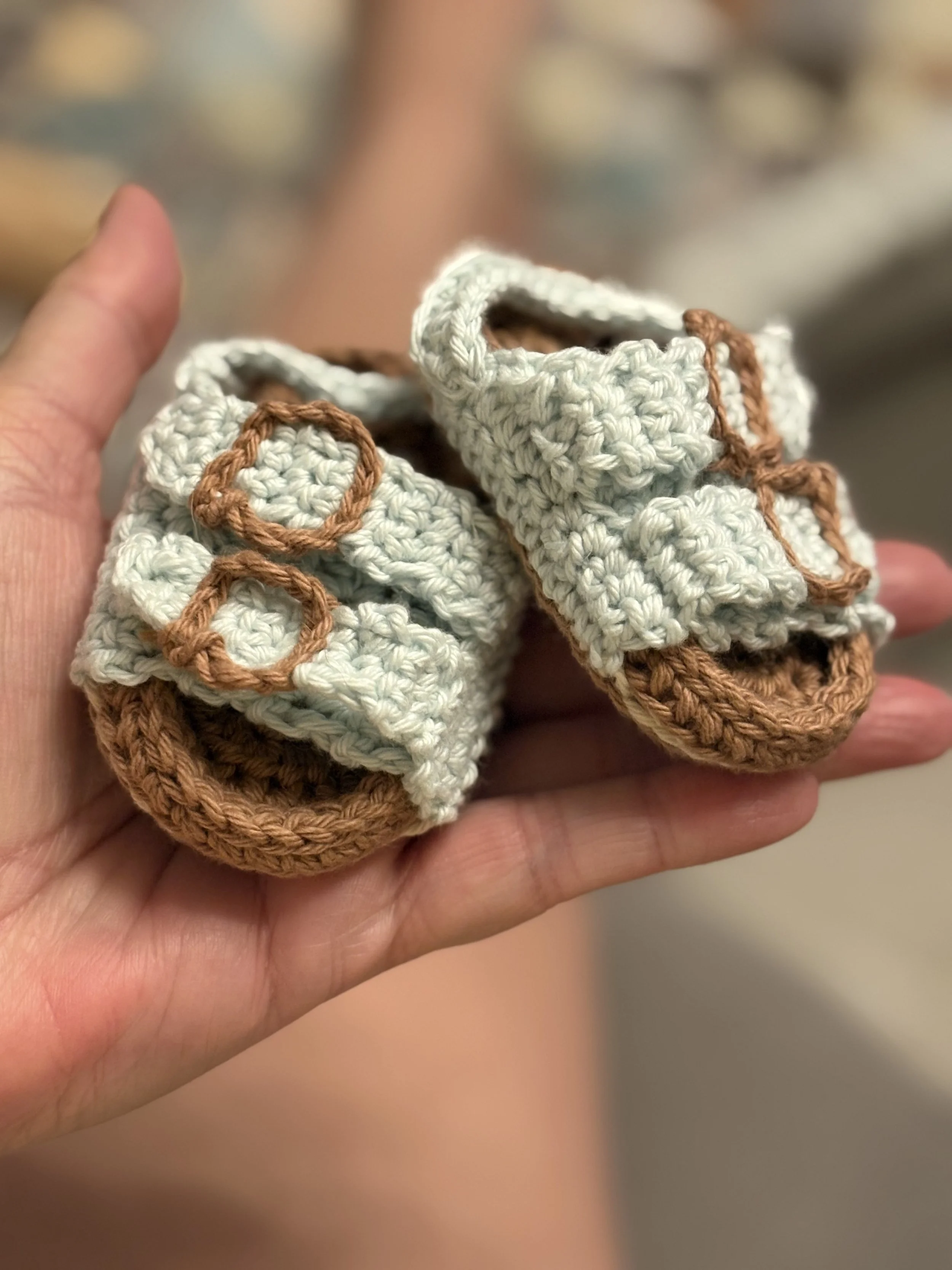 Hand holding small, crocheted baby sandals with brown soles and cream-colored uppers, featuring brown crocheted buckle details