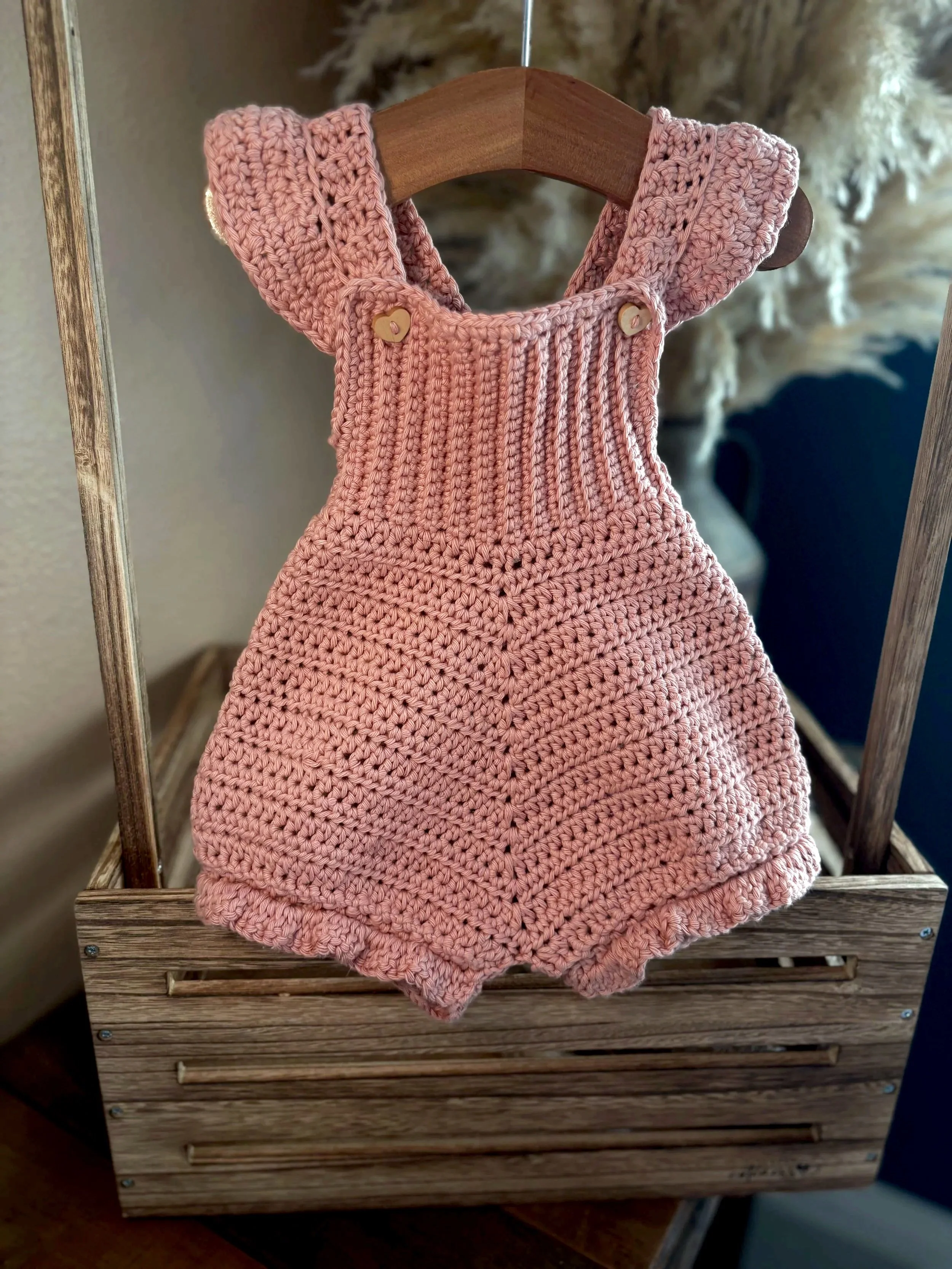Pink crocheted romper with ruffles on shoulders hanging on a wooden clothes rack.