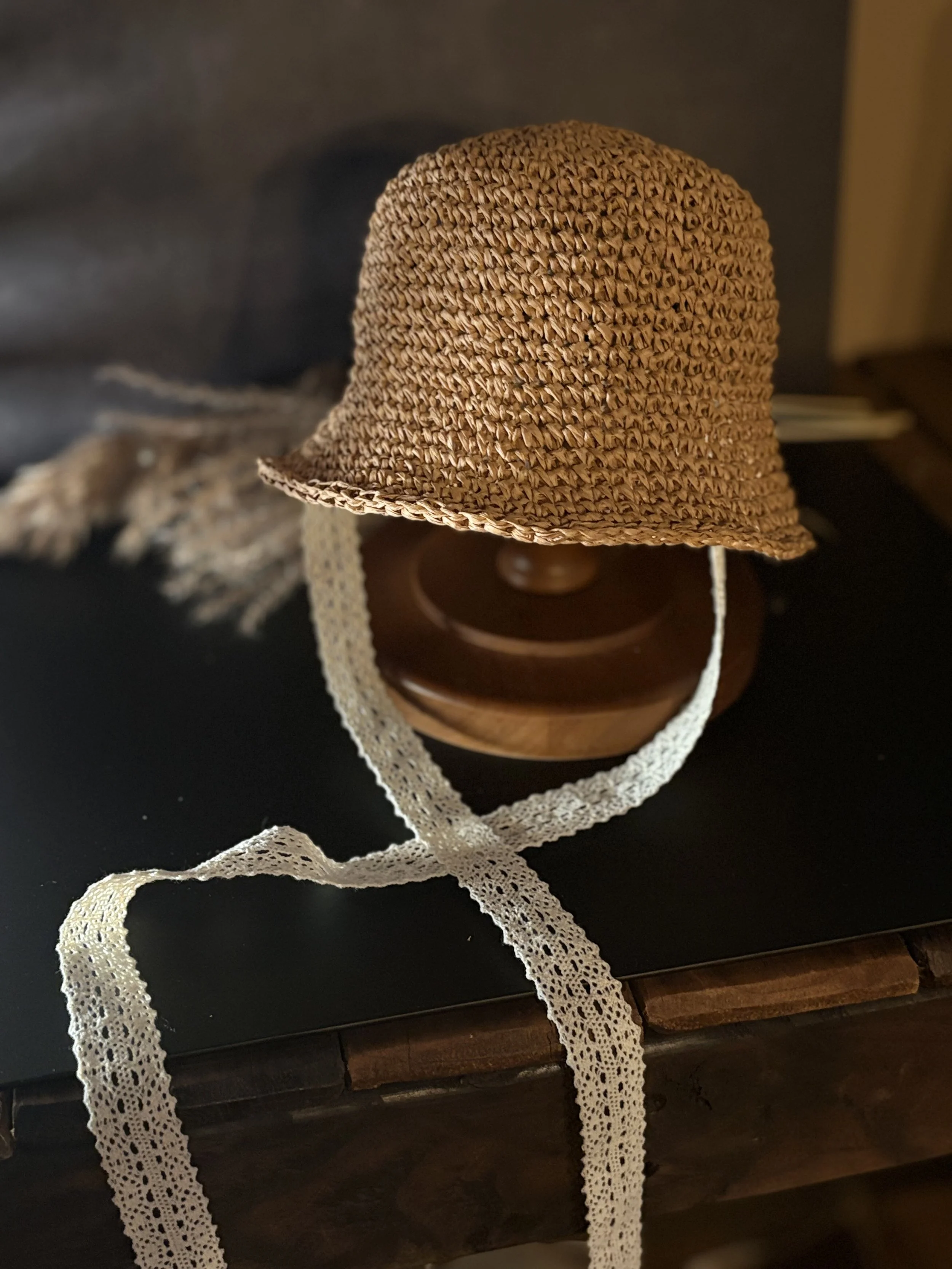 A woven straw children’s hat with a delicate lace tie, resting on a hat stand with a wooden table underneath.