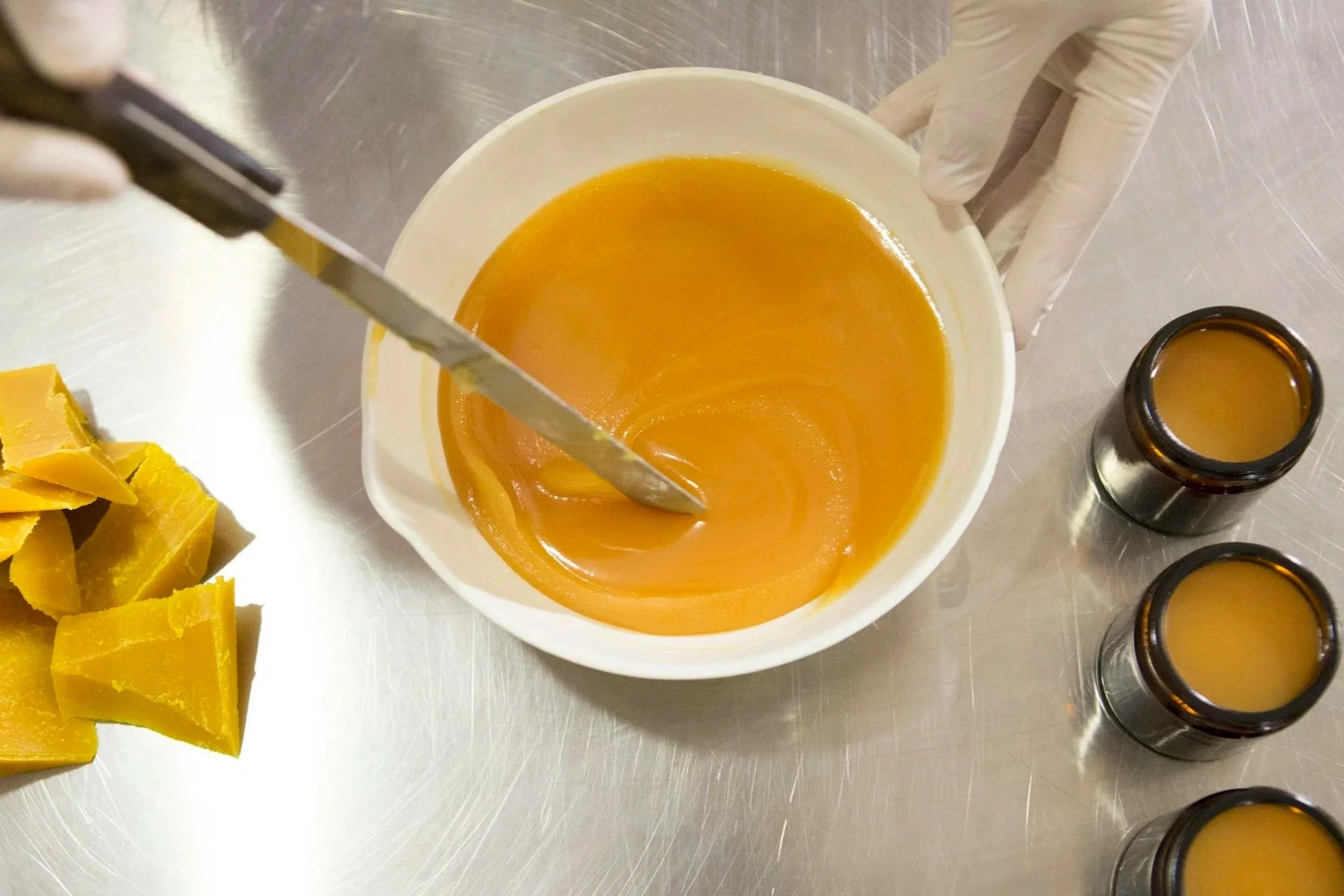 Person wearing gloves stirring oil and beeswax in a white bowl, with small jars of the same substance and a pile of solid beeswax nearby.