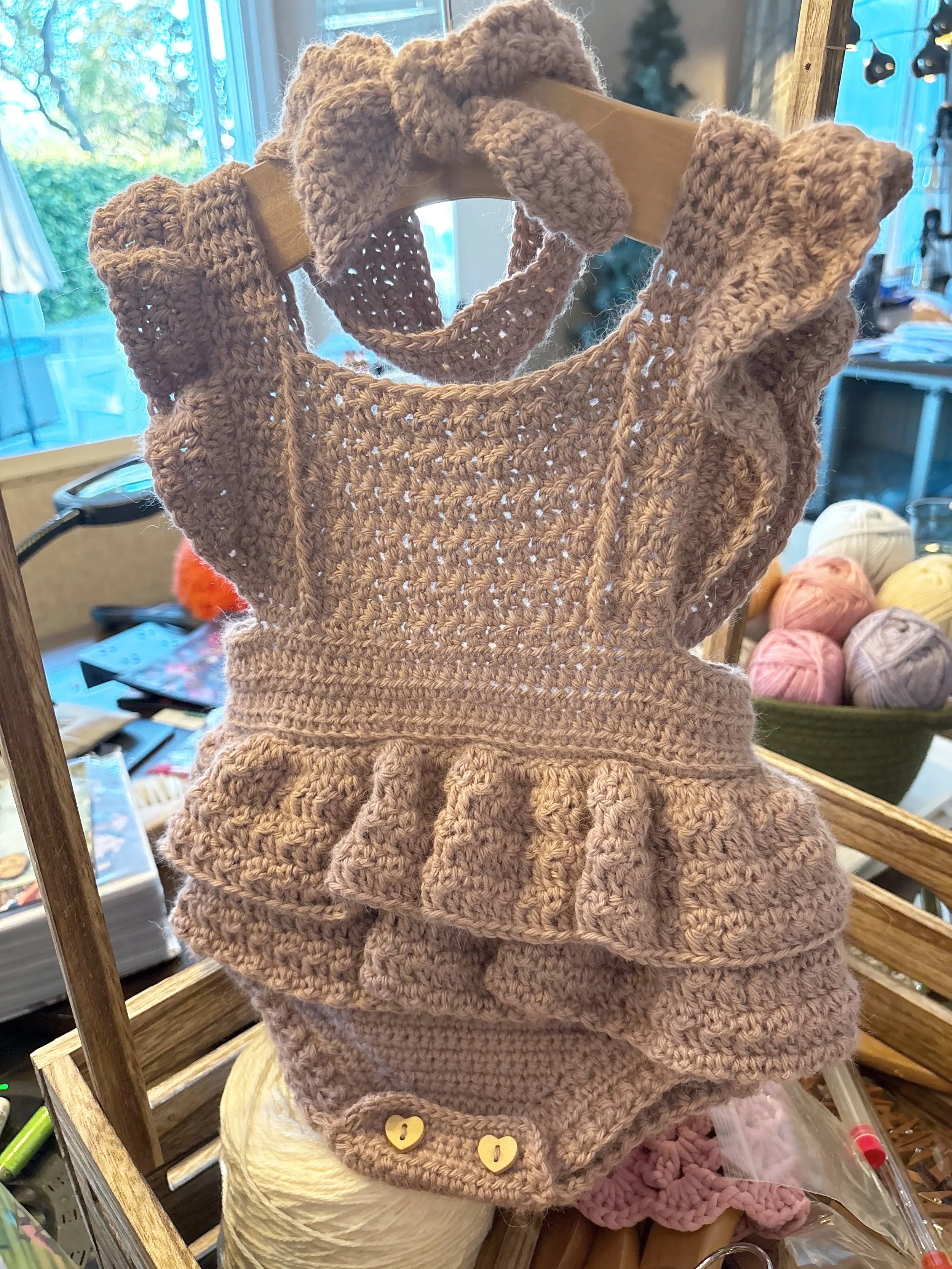A display of a crocheted dusty pink romper with a ruffled hem, buttons made from cutout wood hearts, placed on a mannequin or stand in a craft or yarn store with balls of yarn and craft supplies visible in the background.