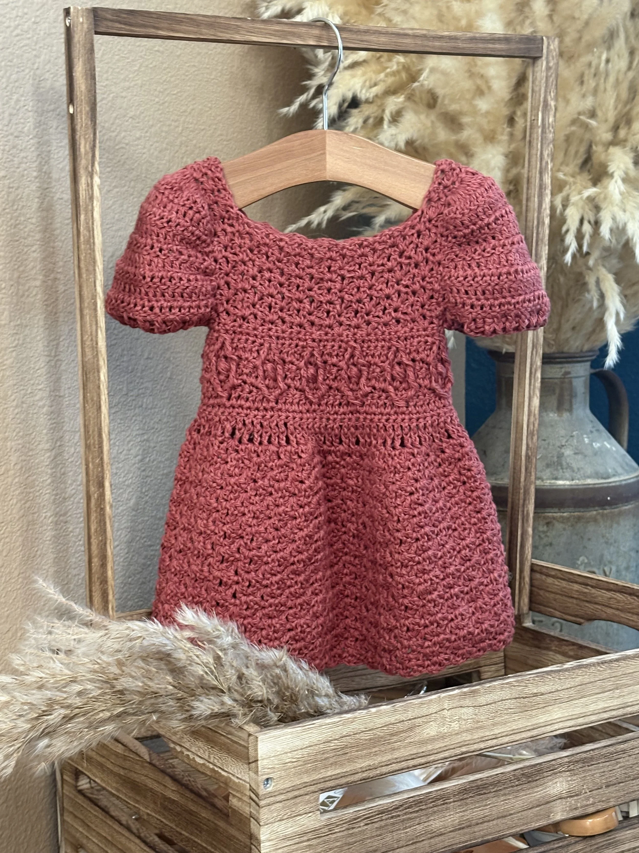 A handmade berry-colored crochet dress for a baby or toddler hanging on a wooden hanger in a rustic setting with dried pampas grass and a metal vase in the background.