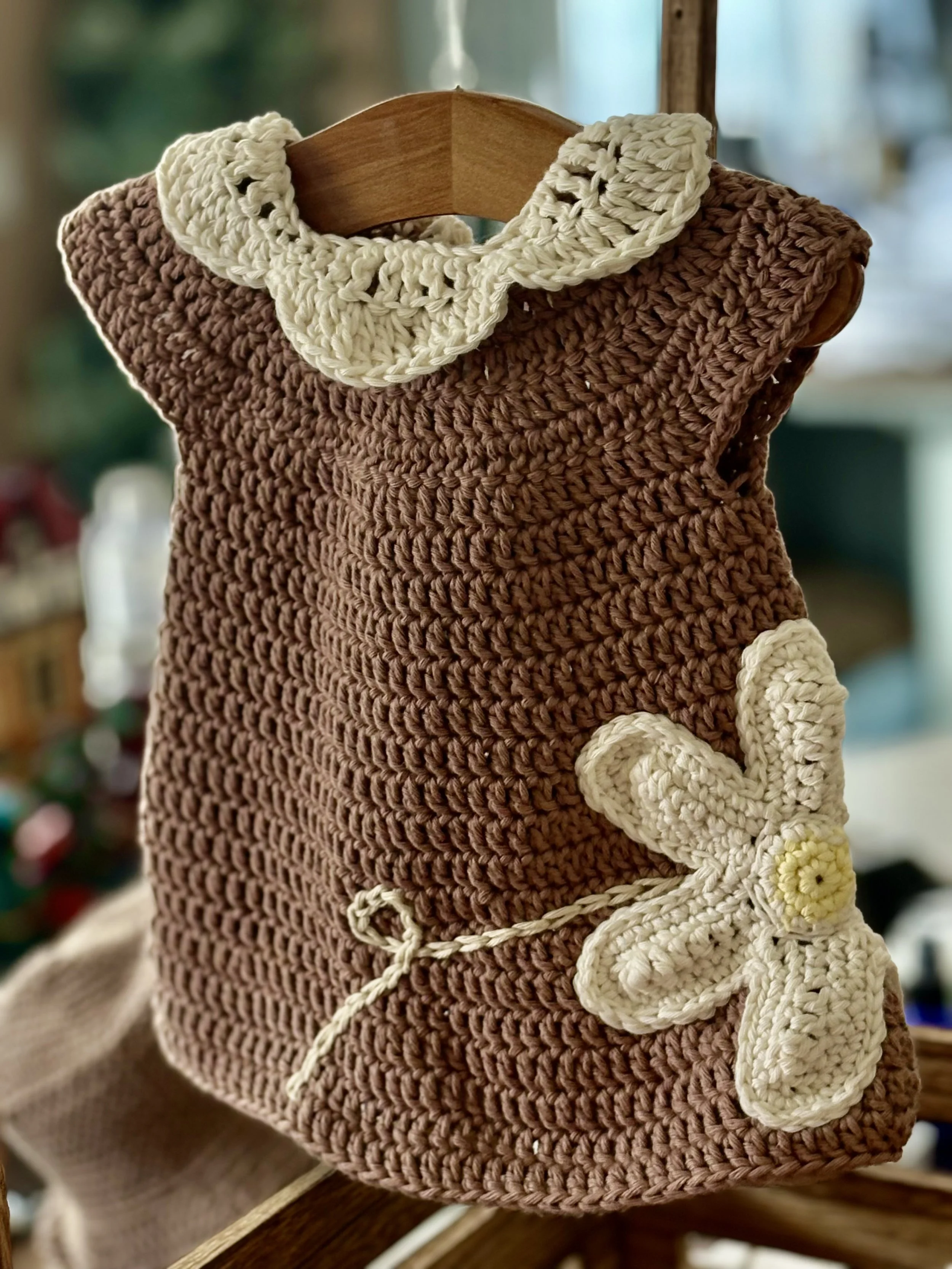 A crocheted summer dress with detailed lace collar in brown with a white flower design, hanging on a wooden display.