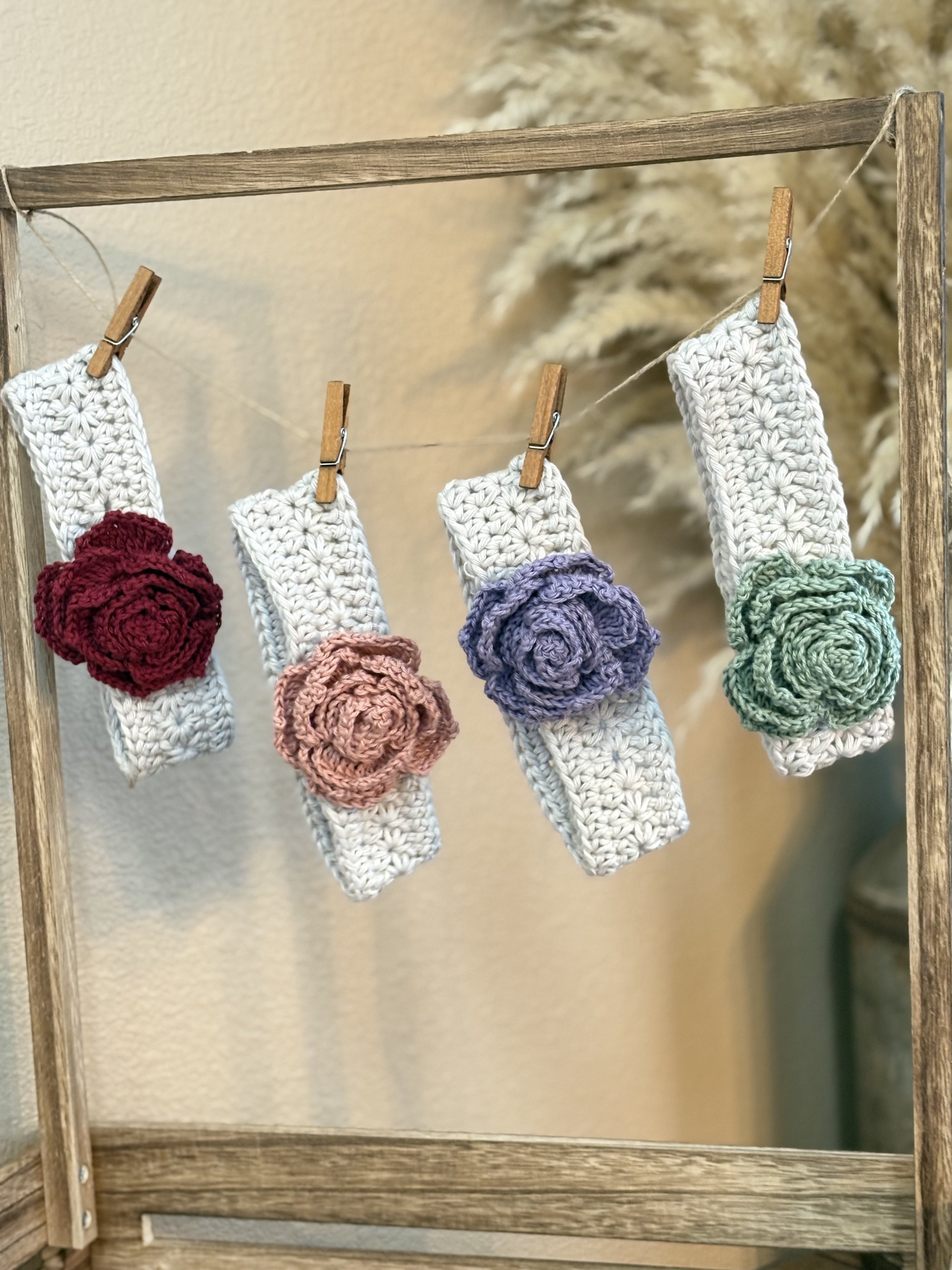 Hand-crocheted headbands for baby, each with a large colorful crochet flower in red, pink, purple, and green, hanging on a wooden frame with clothespins.
