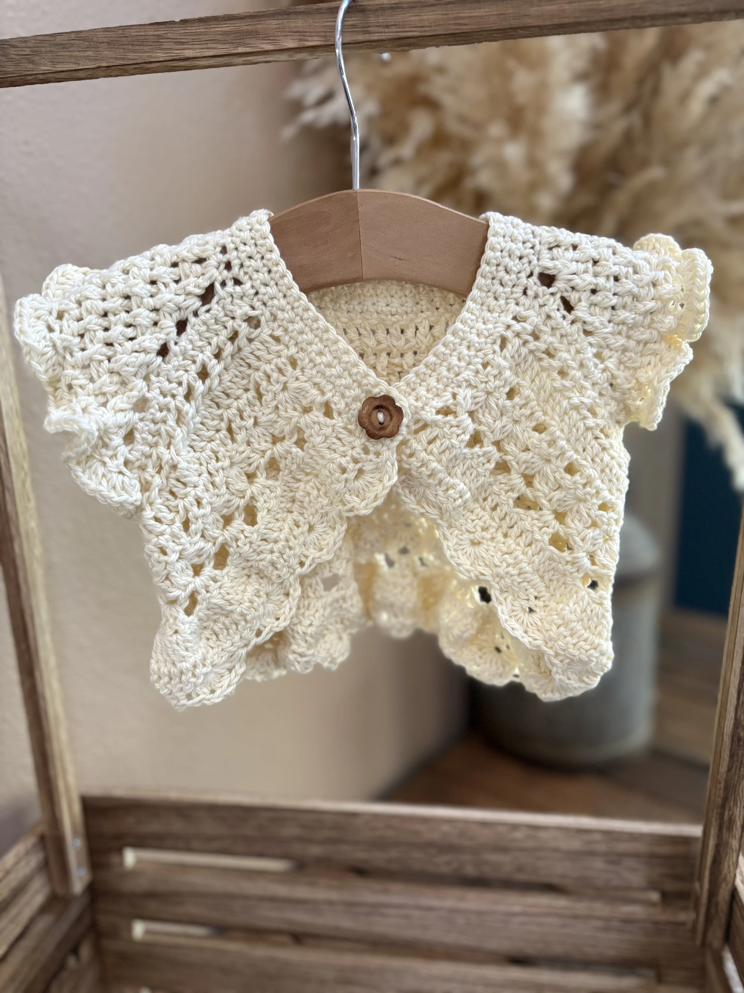 A cream-colored crocheted garment with a button is hanging on a wooden hanger, displayed on a wooden stand.