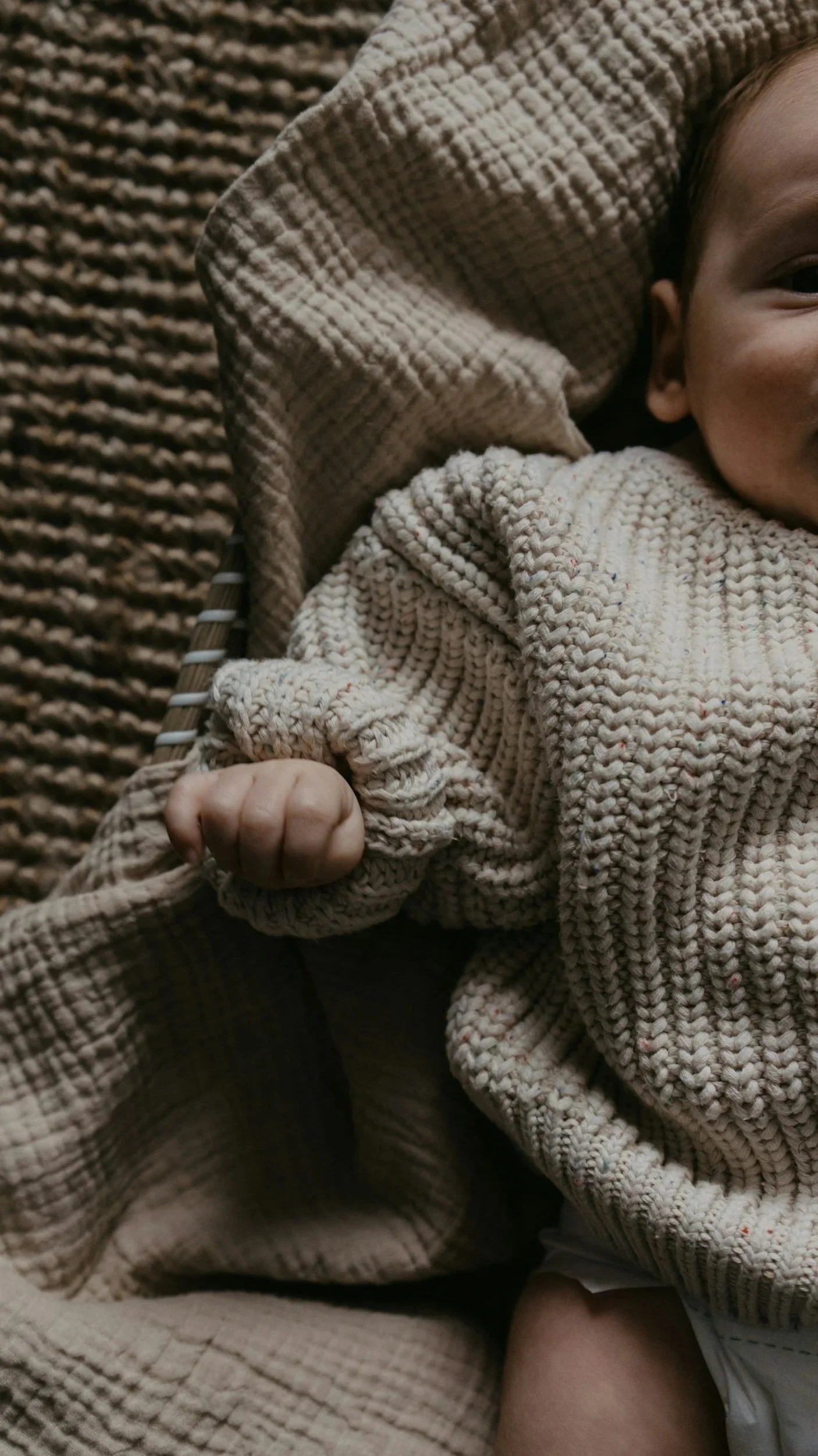 Close-up of a baby lying on a textured brown blanket, wearing a beige crocheted sweater, with a portion of their face visible and their fist clenched.