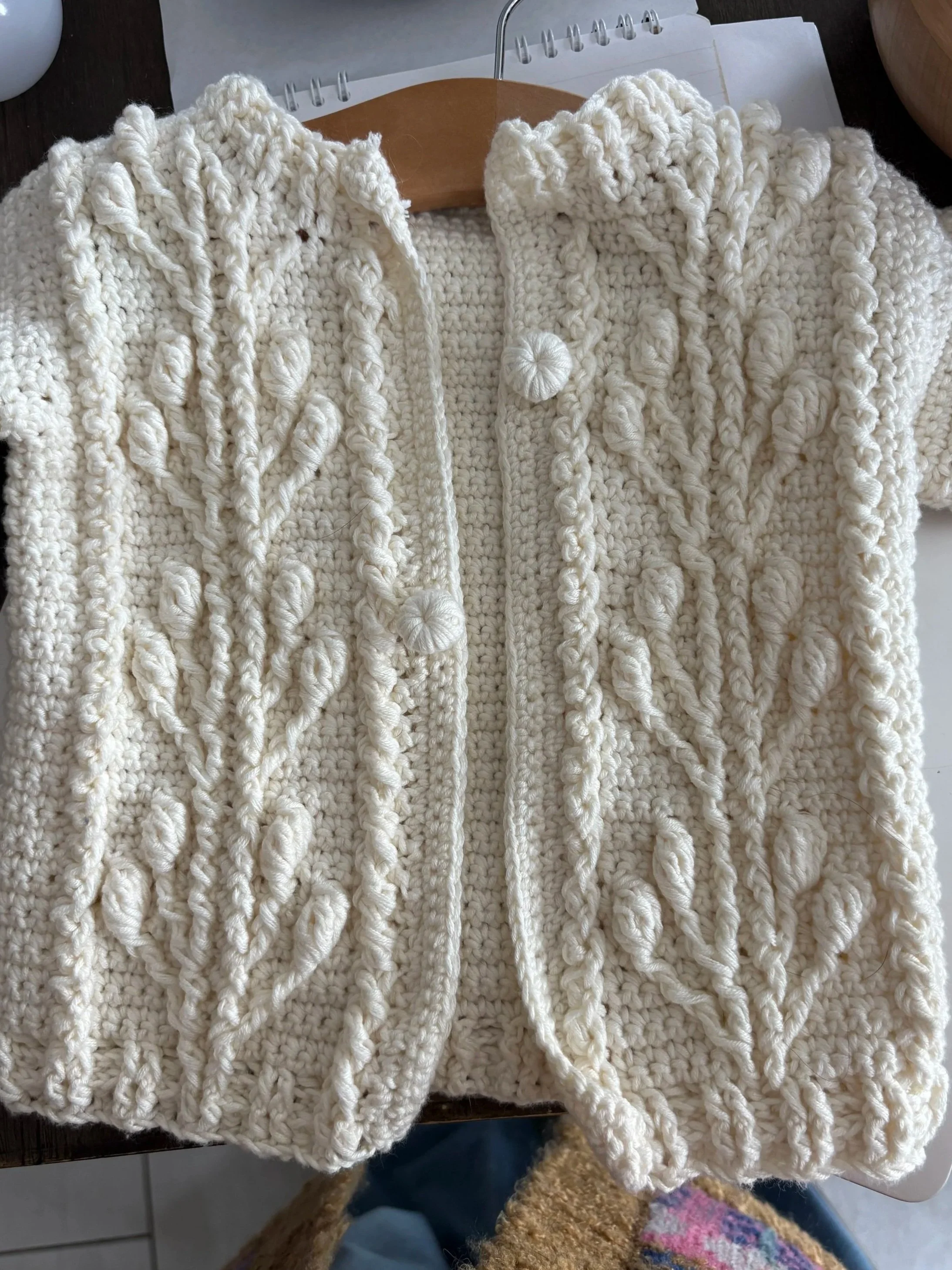 Close-up of a white, hand-crocheted sweater with an intricate botanical and cable pattern, hanging on a wooden hanger.